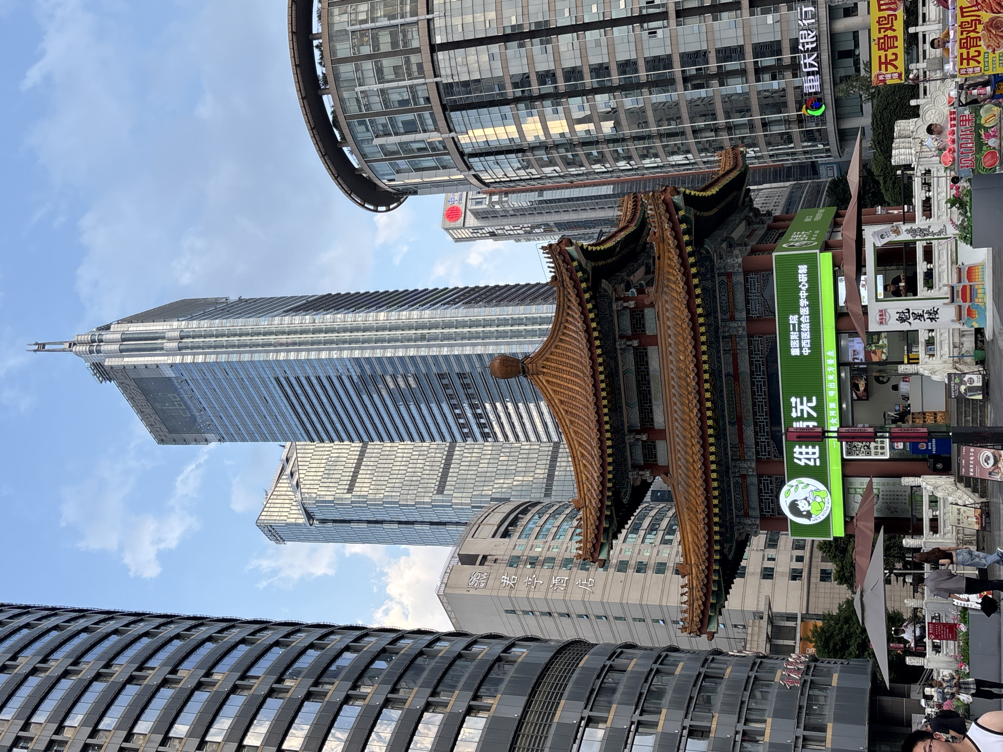 This image depicts a street scene in the Yuzhong District of Chongqing, China, characterized by a juxtaposition of traditional Chinese architecture and modern skyscrapers. In the foreground, a multi-tiered, ornate red-roofed structure, resembling a traditional pavilion or temple, stands prominently. This structure houses a food stall or small restaurant, with a green illuminated sign featuring a cartoon character and Chinese text. Below the pavilion, there is a small plaza area with people walking and vendors selling goods.
Surrounding this traditional element are several imposing modern buildings. To the left, a tall, dark, cylindrical skyscraper with a façade of numerous windows dominates the scene. To the right, a curved, multi-story building with a glass exterior reflects the surrounding environment. Further in the background, other skyscrapers of varying heights and architectural styles pierce the sky. The sky is partly cloudy, with a mix of blue and white.
The image captures a blend of old and new, with the traditional pavilion acting as a focal point amidst the towering contemporary structures, suggesting a dynamic urban landscape. The presence of people, signage, and what appear to be commercial establishments indicates an active public space within the city.