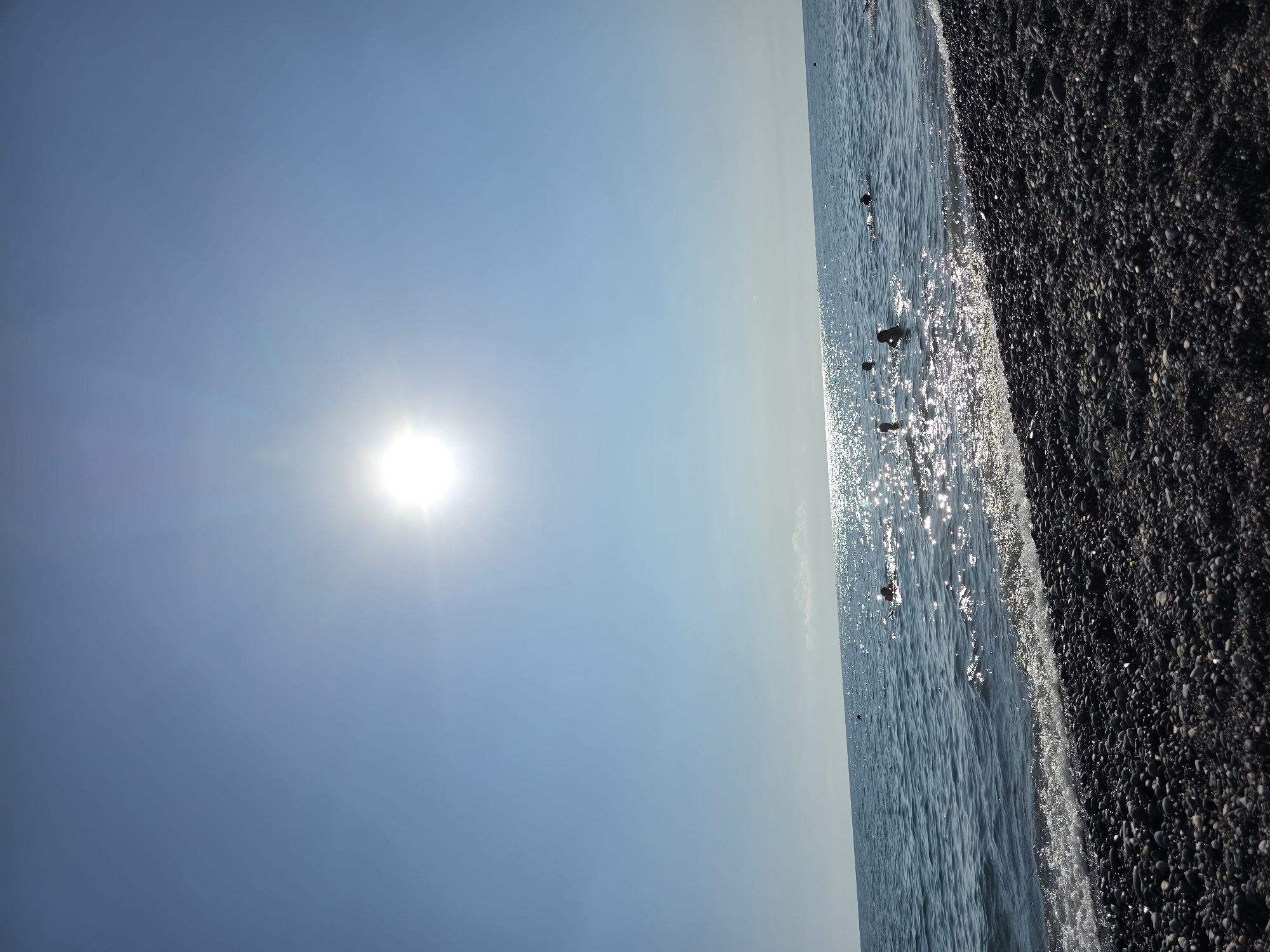 The image captures a tranquil seaside scene at Khostinskiy, Russia. The sun, a brilliant orb in a clear, vast blue sky, bathes the landscape in bright daylight. The sun's rays create a shimmering, sparkling effect on the surface of the calm, blue sea, hinting at a warm and clear day. The sea gently laps against a pebble beach composed of dark, rounded stones of varying sizes. The horizon is a faint, soft line where the sky meets the sea, suggesting a wide, open expanse. A few dark, indistinct shapes appear to be floating on the water, possibly buoys or small boats. The image conveys a sense of peacefulness and the serene beauty of a sunny day by the coast. There is no visible text in the image.