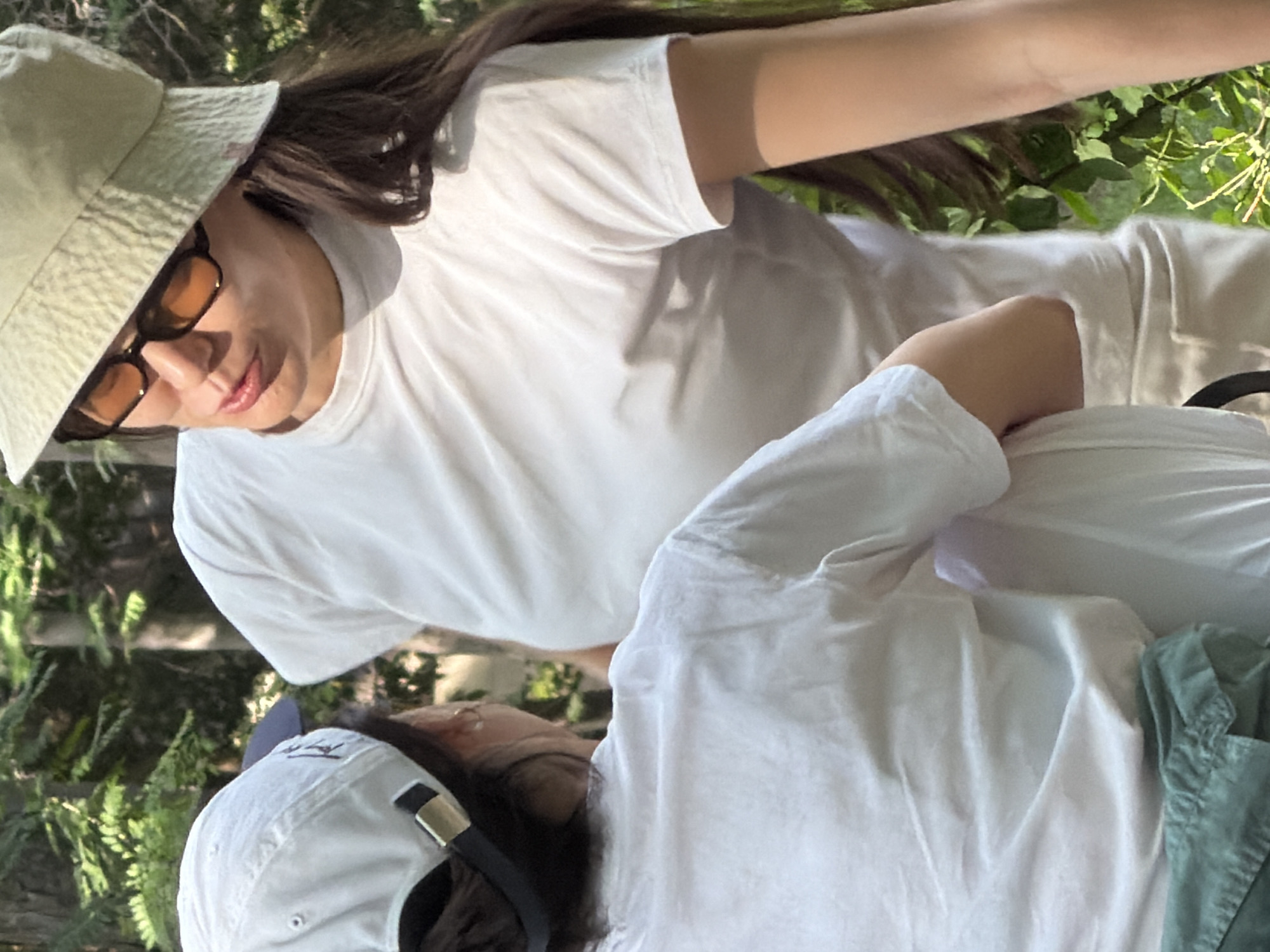 The image features two people, partially visible, wearing white t-shirts and hats. One person, on the left, is wearing a light-colored bucket hat and sunglasses, looking downwards with a slight smile. Their dark hair is visible. The other person, on the right, is wearing a white baseball cap and their face is mostly obscured, but their head is turned down as if looking at something in their lap. The scene appears to be outdoors, with greenery in the background, suggesting a daytime setting. The lighting indicates it is likely sunny. One person's arm is resting on the other's shoulder, hinting at a casual or intimate interaction. There are no visible signs or text in the image.