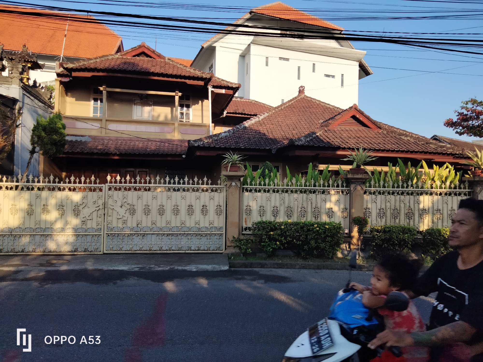 A man is riding a motorcycle with a child passenger on a street in Denpasar, Indonesia, with a gated property featuring a traditional Balinese house and a modern white building in the background. The man, wearing a black t-shirt with a white design, is looking to the right. The child, wearing a red shirt, is seated in front of him and looking to the left. The motorcycle's license plate is partially visible, reading "B 2590 ADH". The street is paved and wet, with reflections of the surroundings. The property is enclosed by an ornate white metal gate and a low hedge. The architecture of the buildings suggests a blend of traditional and modern styles, with terracotta tile roofs and wooden accents on the older structure, and a stark white facade on the newer one. Power lines are visible crisscrossing the clear blue sky. The scene appears to be captured during daylight, possibly late afternoon, given the warm lighting and shadows. The OPPO A53 watermark is present in the bottom left corner.