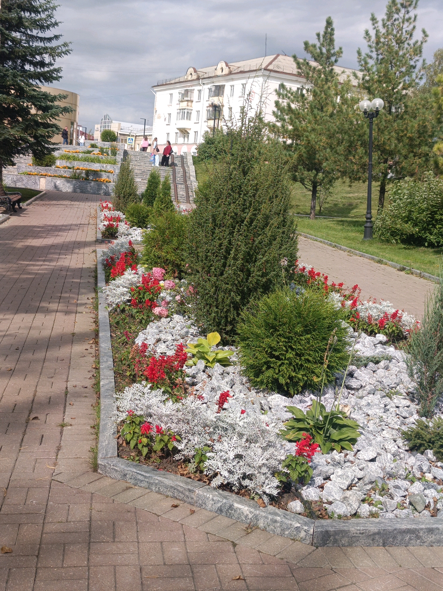 The image depicts a paved pathway in a park-like setting in Satka, Russia, with a tiered flowerbed bordering the walkway on the right. The flowerbed features a variety of plants, including lush green bushes, ornamental grasses with silvery foliage, and bright red flowers. The pathway is constructed of rectangular brick pavers. In the background, a multi-story building with white walls and multiple windows is visible, suggesting an urban or semi-urban environment. Several people are present on a set of stairs leading up towards the building. The sky is overcast with scattered clouds.