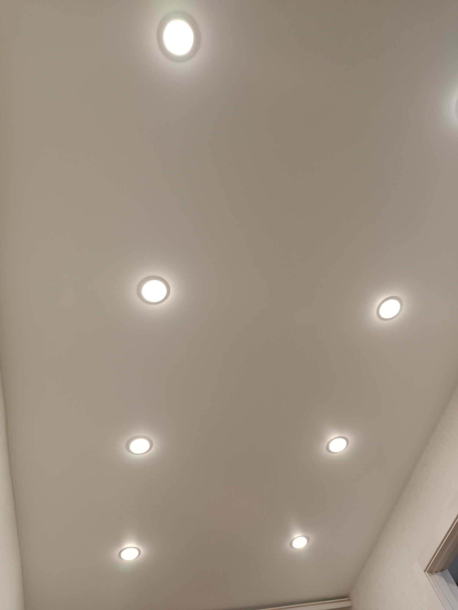 The image shows a ceiling with recessed lighting. There are eight round lights evenly spaced in a rectangular grid pattern. The ceiling is off-white. In the lower right corner, a section of wall and window trim is visible. The location is uncertain, but the mention of the city of Chaikovsky, Russia, might suggest it is an interior space in that city.