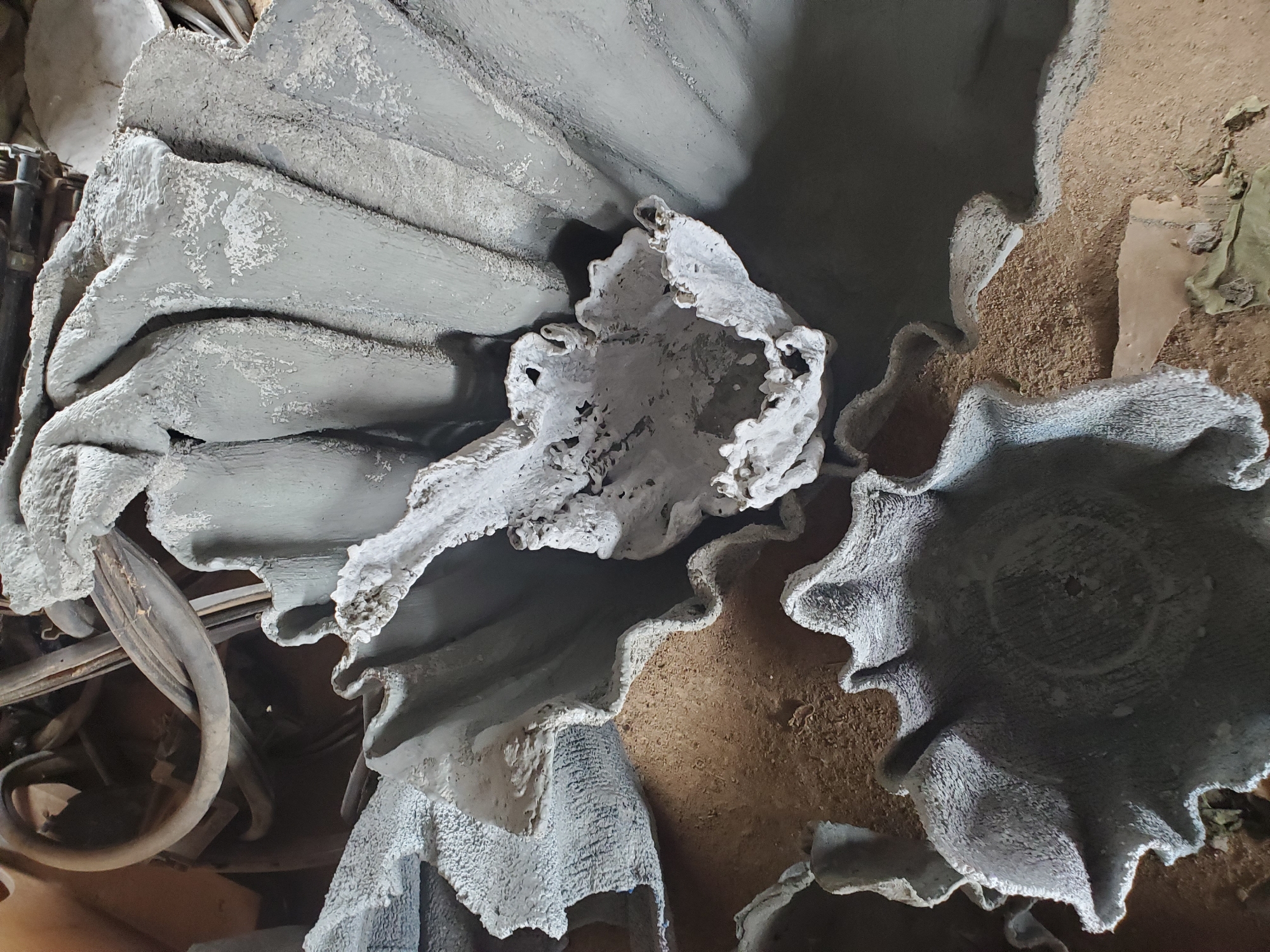 A close-up, overhead shot captures several large, rough, gray concrete planters in an outdoor, possibly workshop, environment. The planters have irregular, wavy edges, resembling petals or large leaves. Some are partially filled with a sandy, dirt-like material. In the background, out of focus, are various discarded items including coiled electrical cables and what appear to be metal components. The lighting is harsh, creating strong shadows that emphasize the textured surfaces of the planters. The scene suggests a craft or construction area, possibly in Jos, Nigeria, where these planters are being made or stored. There are no people or animals visible in the image.