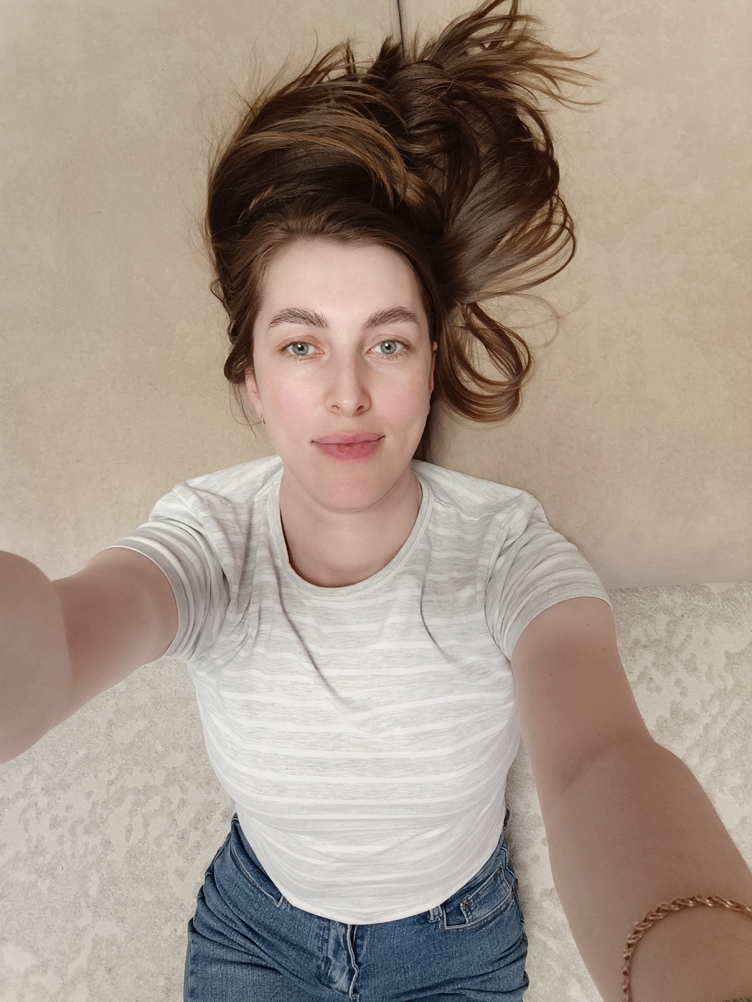 The image is a high-angle selfie of a woman lying down indoors. The subject is a woman with fair skin, blue-green eyes, and long brown hair fanned out around her head. She looks directly into the camera with a gentle, soft smile, conveying a relaxed and pleasant expression.
She is dressed casually in a light-colored, possibly white or light grey, short-sleeved t-shirt with subtle horizontal stripes, paired with blue denim jeans. A golden-colored, braided bracelet or chain is visible on her right wrist, which is extended upwards as she holds the camera to take the photo. Her left arm is also extended, partially visible on the left side of the frame.
The background behind her head is a plain, light beige or off-white surface, which could be a floor, bed, or sofa. The lower portion of the image, beneath her body, shows a textured light-colored rug or carpet with a subtle pattern. The setting appears to be a comfortable indoor space, likely a home. The lighting is soft and even, without strong shadows, making it difficult to determine the specific time of day. There is no visible text in the image.