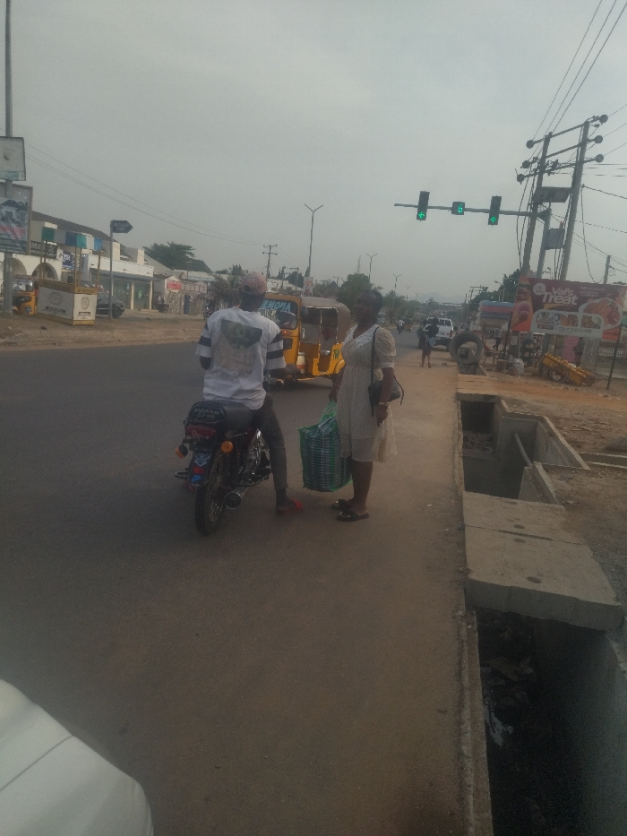 The image depicts a scene on a road in New Karu, Nigeria. A man wearing a white t-shirt and a cap is seated on a black motorcycle. The motorcycle is facing left and its engine is running. Behind the man is a woman in a long, white dress holding a green and white shopping bag. The man is looking towards the woman who is looking directly at the camera. Behind them, a yellow auto-rickshaw is visible, as are several buildings on the left side of the image. In the background, there is a set of three traffic lights, the middle one of which is green. To the right of the woman, there is a concrete drainage ditch, and a sign for a fast-food restaurant. There are several trees and bushes along the edge of the road and sidewalk. The sky is overcast and the lighting is diffuse. The scene is quiet and calm. The people appear to be waiting for the motorcycle to move.