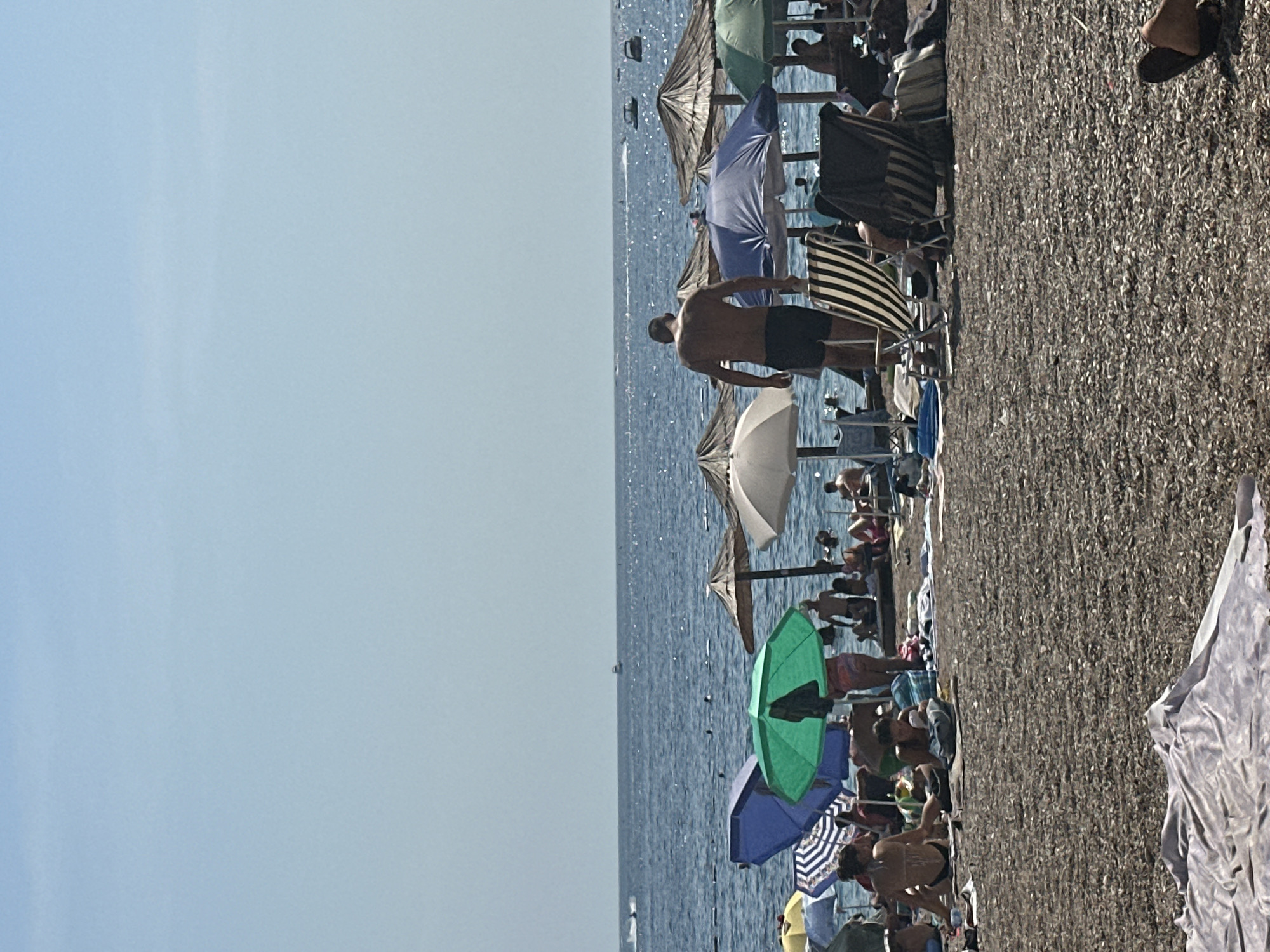 The image depicts a crowded beach scene in Budva, Montenegro, on a sunny day. The foreground is filled with pebbles and a light-colored cloth. Beyond this, numerous beach umbrellas, primarily straw and some fabric in blue and green, are scattered across the sand, providing shade for beachgoers. People are seen sitting, lounging on chairs, and walking on the beach. In the midground, a man wearing black shorts walks away from the camera, facing the sea. Several beach chairs, some striped and some dark, are visible. The sea, a deep blue, stretches to the horizon, with a clear, pale blue sky above. A few boats and jet skis are visible on the water in the distance. The overall atmosphere is that of a typical summer day at a popular tourist destination.
