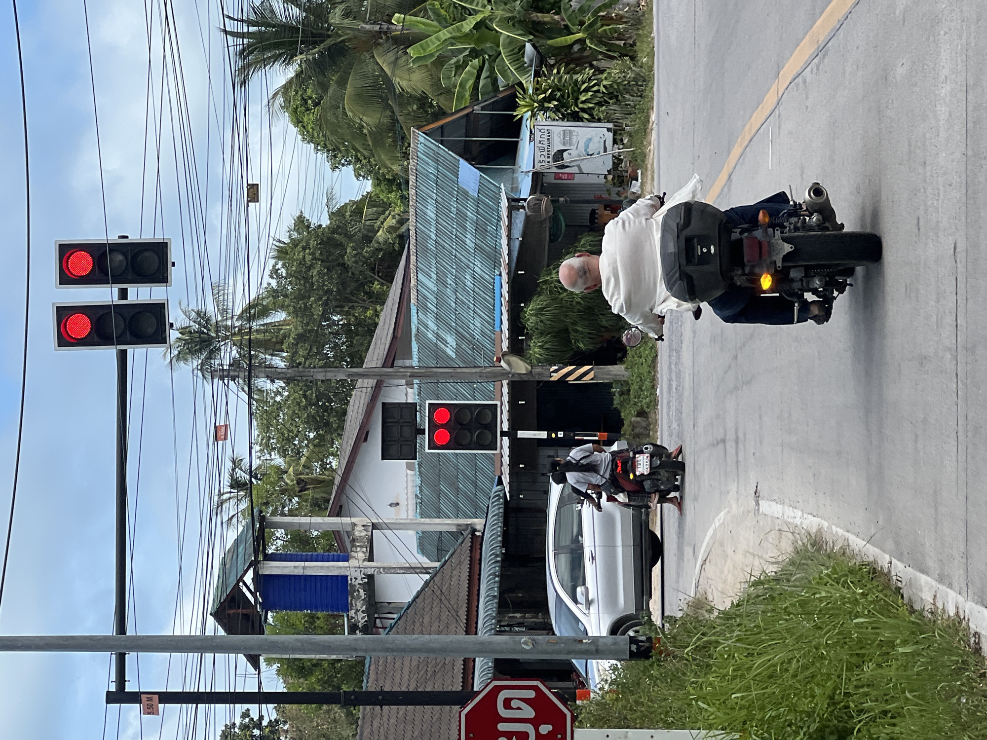The image shows a street scene in Tambon Ko Phangan, Thailand. Two motorcycles are visible. The first motorcycle, in the foreground, is ridden by a man wearing a white shirt and dark pants. He has a black storage box on the back of his motorcycle, which is facing away from the camera. The second motorcycle is further down the road and is carrying two people. A silver SUV is parked on the left side of the road. A traffic light with red lights illuminated is visible above the second motorcycle. A red octagonal stop sign is also present on the left side of the image. Buildings with corrugated metal roofs are present in the background, along with palm trees and utility wires. The sky is partly cloudy.