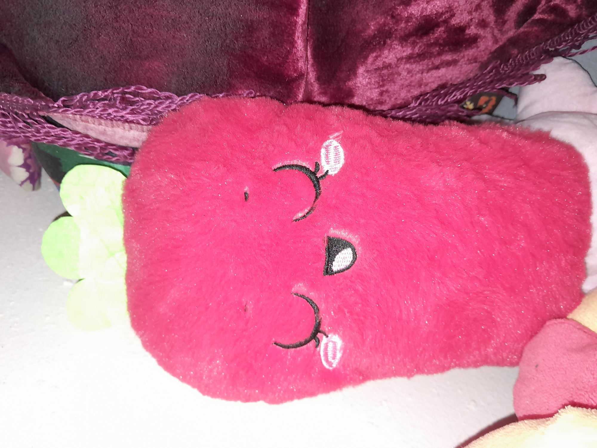 The image shows a close-up of a plush toy resembling a pink radish with a cute face, featuring closed eyes with eyelashes and a smiling mouth. The toy has green leaves at the top. It is positioned next to other plush toys and fabrics, one of which is a deep purple velvet. The background is a plain white wall on the left. There is no visible indication or context that places this image in the city of Uchquduq, Uzbekistan, or any other specific geographic location.