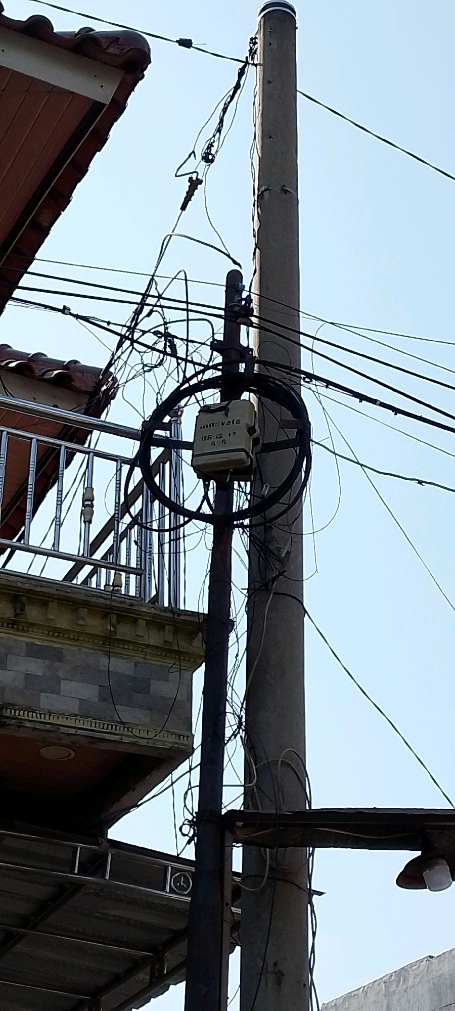 The image captures a utility pole with numerous tangled wires and a beige electrical box. A portion of a building with a tiled roof and a metal railing is visible to the left. The sky is clear and blue. The scene appears to be in a residential or commercial area, likely in Satriamekar, Indonesia, given the context. There are no people or animals visible in the frame. The overall impression is one of cluttered but functional infrastructure.