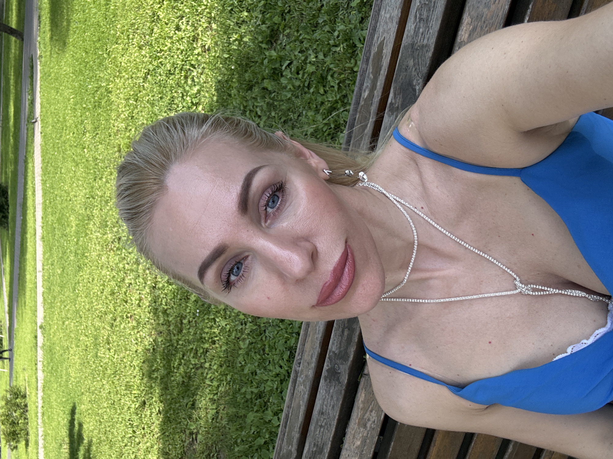 The image is an upward-angled selfie of a woman taken outdoors on a sunny day, likely in a park or garden. The woman, appearing to be in her 30s or 40s, has blonde hair, striking light blue eyes, and fair skin. She looks directly into the camera with a calm expression, her lips adorned with rosy-mauve lipstick. She wears a vibrant royal blue strap top or dress, with a hint of white lace trim visible along her neckline. A distinctive silver-toned, sparkling necklace wraps around her neck and cascades down her décolletage, featuring an intricate knot detail in the center. Small silver stud earrings are also visible.
She is seated or reclining on a rustic wooden bench, parts of which are visible on the right and bottom of the frame. The background is dominated by lush, bright green grass, indicative of a well-maintained outdoor space. Shadows suggest bright sunlight, placing the scene in daylight, possibly late morning or afternoon. In the distance, a narrow path cuts through the grass, bordered by faint lines, and some small bushes are sparsely scattered. The image conveys a serene outdoor moment. No visible text is present.