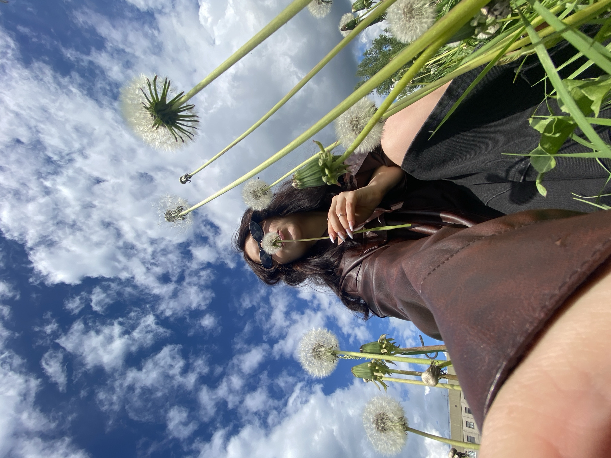 The image shows a woman, identified by her dark hair, sunglasses, and brown leather jacket, holding a dandelion in front of her face. She has manicured nails. She is wearing a black garment. The perspective is from below, looking up at her and a blue sky with puffy white clouds. Numerous dandelions in various stages of seed dispersal surround her. A building can be seen at the bottom. The location is Марковский, Russia.