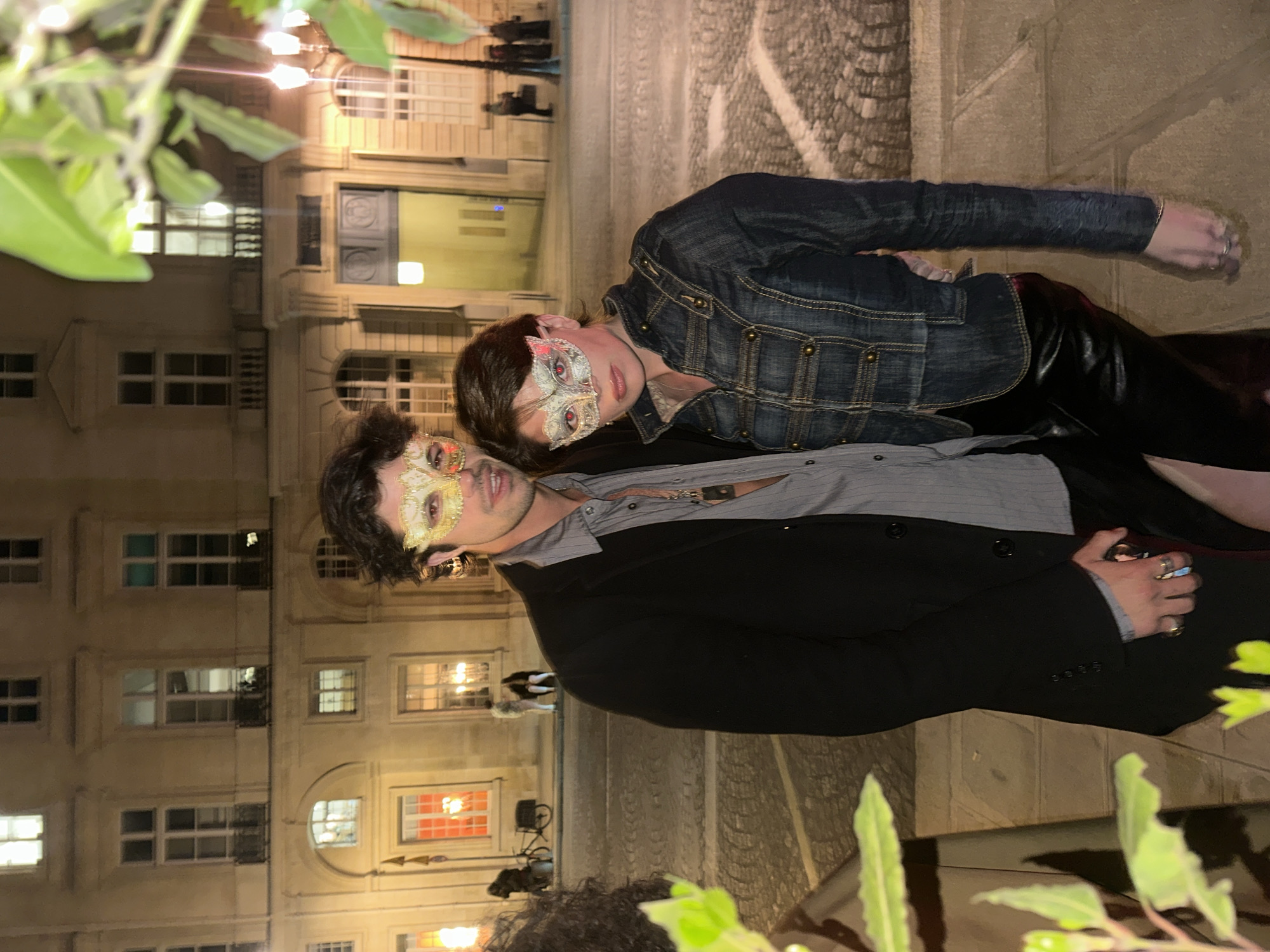 A man and a woman are posing for a photo on a street in Paris, France. The man is wearing a black suit jacket over a grey striped shirt, and a gold masquerade mask. The woman is wearing a denim jacket over a black top, and a silver masquerade mask. They are standing close together and looking at the camera. The background consists of a building with lighted windows. The street appears to be cobblestone. There are plants visible in the foreground. The lighting is dim.