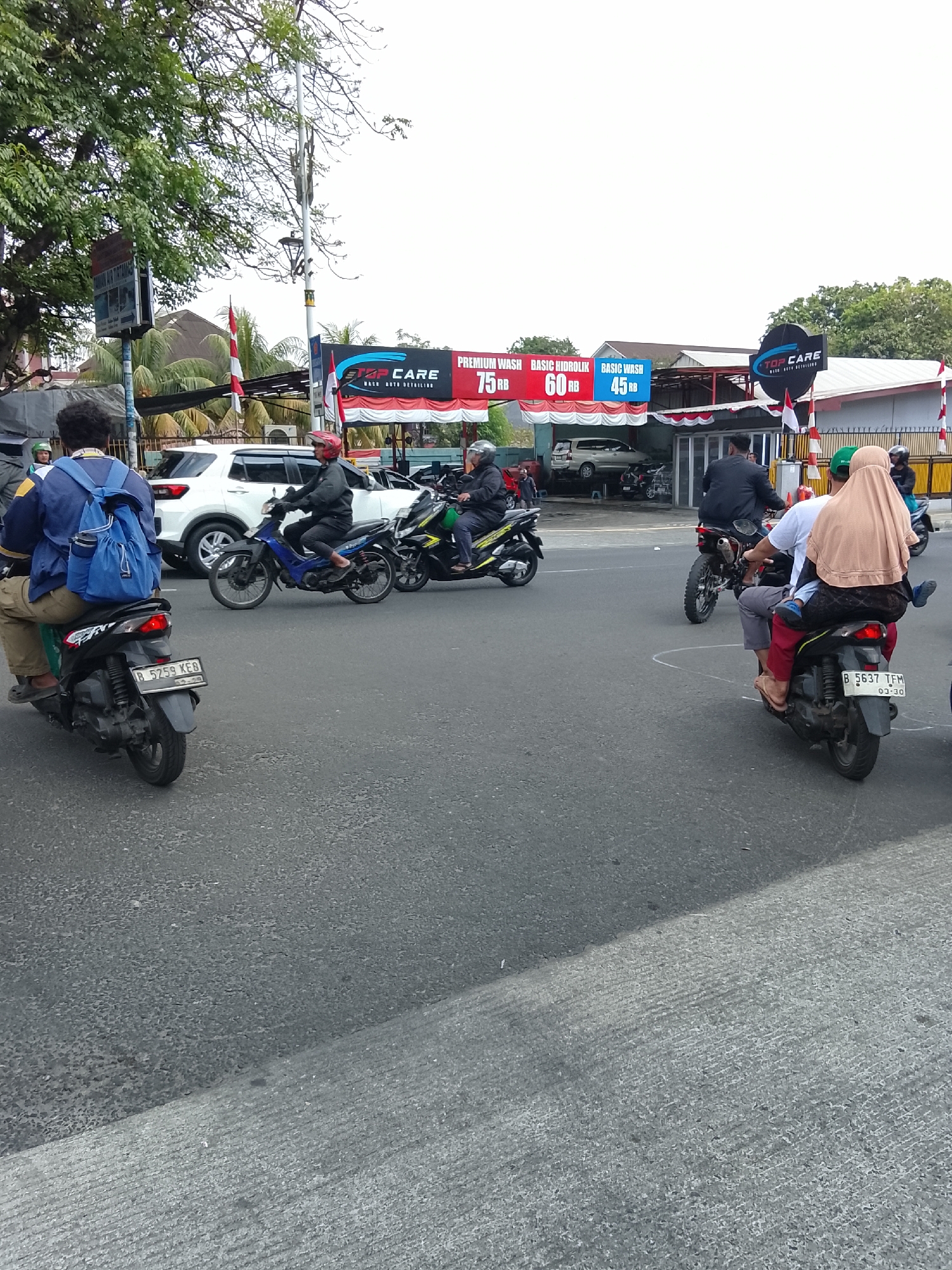 The image captures a street scene in East Jakarta, Indonesia, featuring a car wash establishment in the background and several individuals on motorcycles navigating the road. The car wash, branded "TOP CARE," advertises "PREMIUM WASH," "BASIC HIDROLIK," and "BASIC WASH" with prices listed in Indonesian Rupiah (RB). Red and white flags, possibly the Indonesian flag, are displayed at the entrance of the car wash.
In the foreground, a person wearing a blue backpack rides a black motorcycle with the license plate "B 5259 KEB." To their right, another individual on a blue motorcycle is visible. Further to the right, three people are riding on a black motorcycle; the passenger in the back is wearing a light brown hijab and a black skirt, with a child's legs visible dangling. This motorcycle has the license plate "B 5637 TFM." Another motorcycle, a dirt bike style, is seen behind the blue motorcycle. A white SUV is parked at the car wash. The overall environment depicts a typical urban street in Indonesia, with traffic and commercial activity.