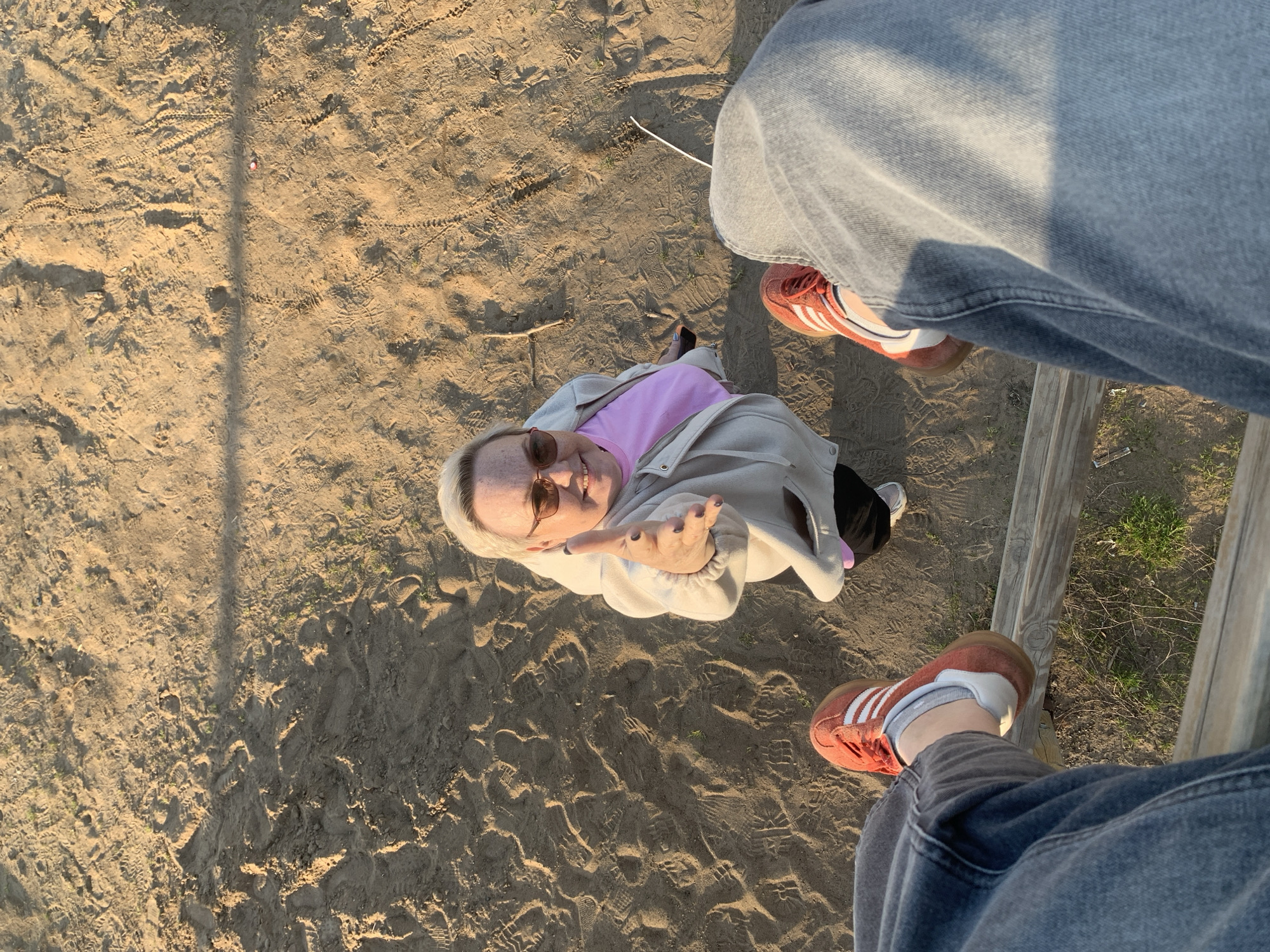 Captured from an elevated perspective, the image portrays a woman on sandy terrain. She is clad in a pink shirt and a light-colored jacket. The photograph includes parts of a wooden structure and a person's legs, with their feet visible in orange and white sneakers. The scene is on the sand of the beach in Chaikovsky, Russia.
