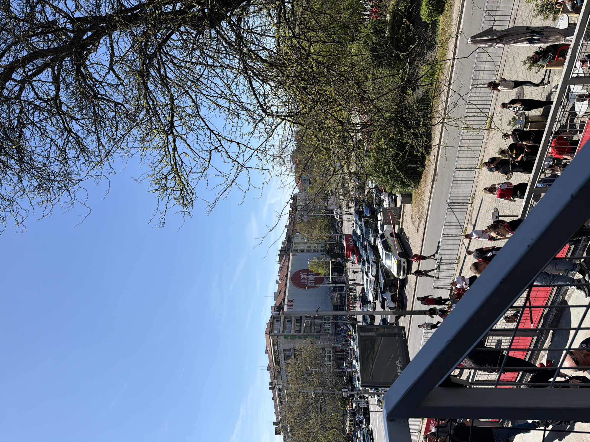A daytime shot from a high vantage point, likely a balcony, presents a street scene in Sofia, Bulgaria. The view, partially framed by bare tree branches, captures a street lined with buildings, parked vehicles, and pedestrians. One building prominently features a large advertisement for "LUCKY STRIKE" and the word "HOBO" below. The street is divided by metal barriers, and a gathering of people is observed on the side of the building and across the barrier. A grey railing of the balcony where the picture was taken is in the foreground on the right. The sky is clear with a few clouds.