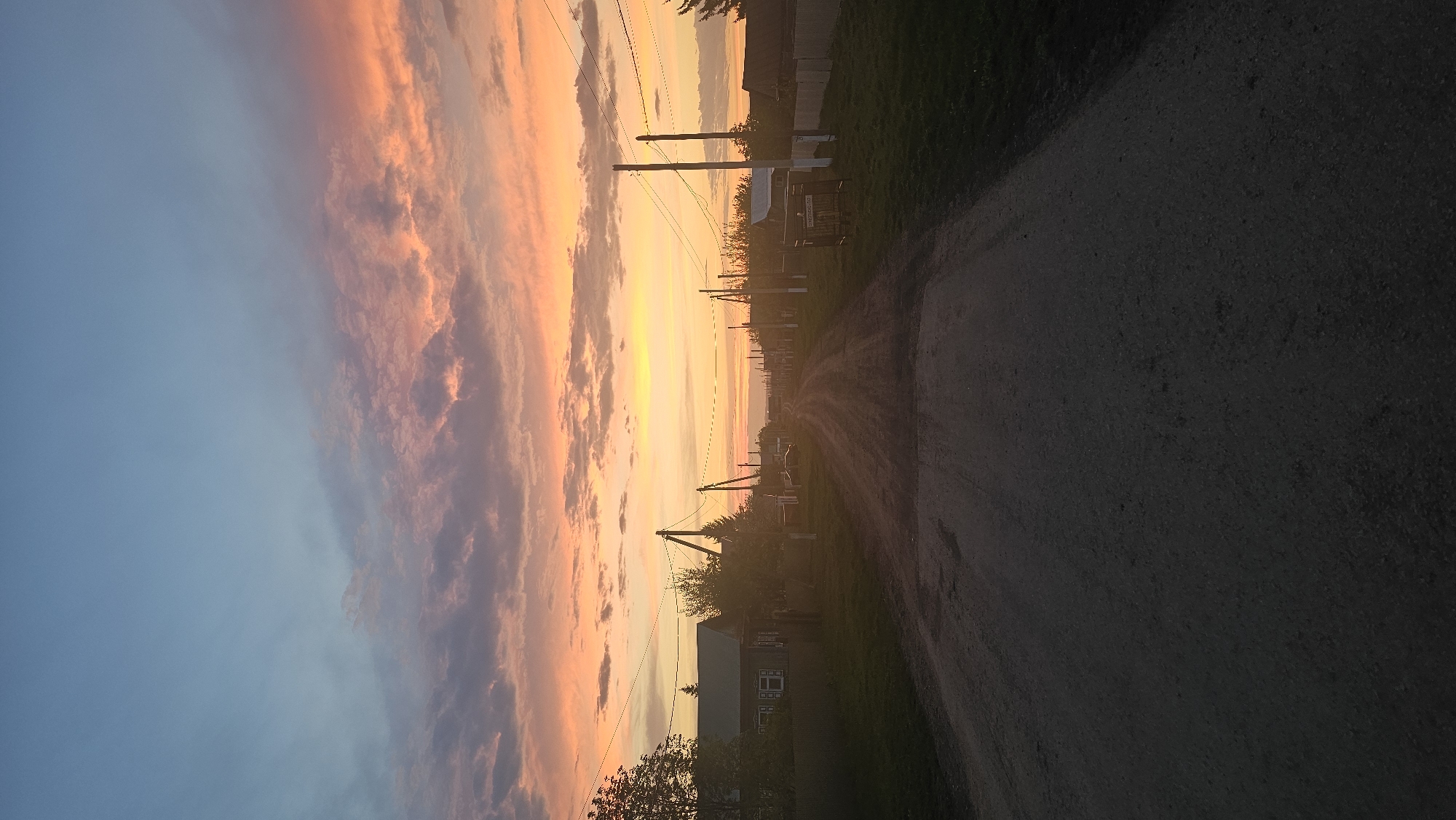 A vertical view captures a rural scene during a vibrant sunset in Yangiskain, Russia. The sky exhibits a gradient of colors from blue to orange, with clouds tinged in shades of orange and pink. A dirt road runs from the foreground into the distance, flanked by utility poles and residential structures. Vegetation lines the road's edge. The time is around sunset.