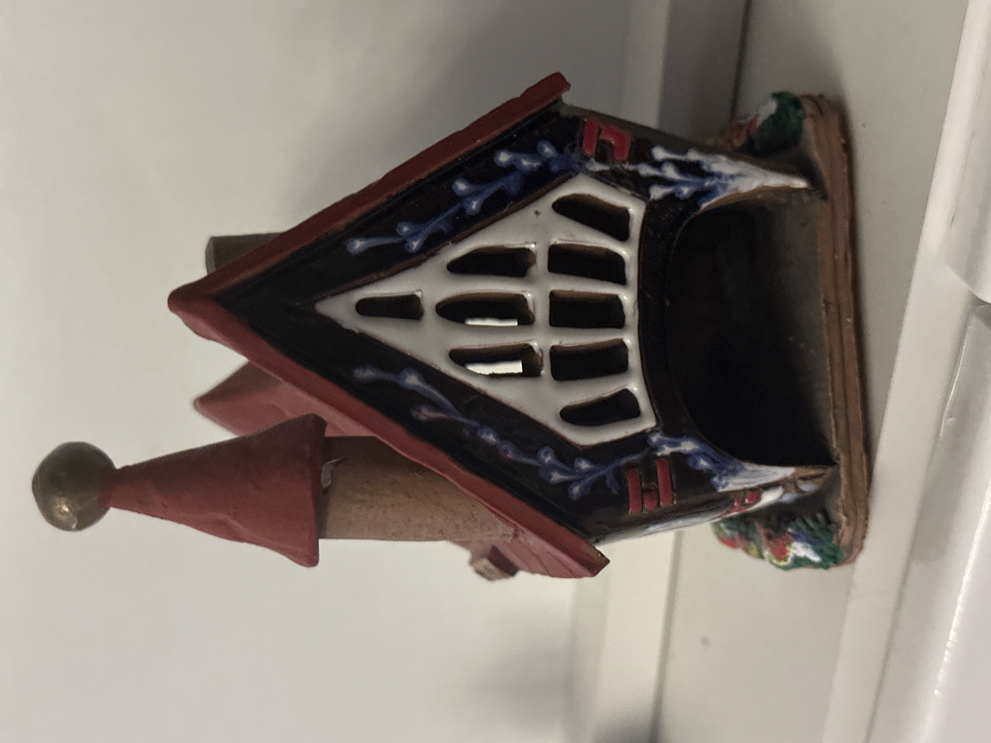 The image displays a decorative, house-shaped object, likely made of ceramic or painted material, placed on a plain, light-colored surface. The object is oriented to its side in the main image but the crops clarify its upright structure.
The miniature house features a prominent red, pointed roof. From one side of the roof, a slender, red conical spire with a small golden or brass ball finial extends upwards. The main body of the house is dark, possibly black or dark brown, and is adorned with intricate white and blue decorative patterns resembling vines or swirls. A large, stylized window-like opening on the front is painted white and features several horizontal bars, giving it a gridded appearance. Small red details, possibly stylized letters or shapes resembling "H", are visible on the dark body near the roofline. The base of the house appears to be a natural, earthy tone with small, colorful accents in green, red, and white, suggesting miniature foliage or floral elements.
No people are visible, and no activity is taking place; the scene is static. The setting appears to be indoors, against a uniform light background. There are no specific visible cues within the image to indicate the provided location context of Bobov Dol, Bulgaria.