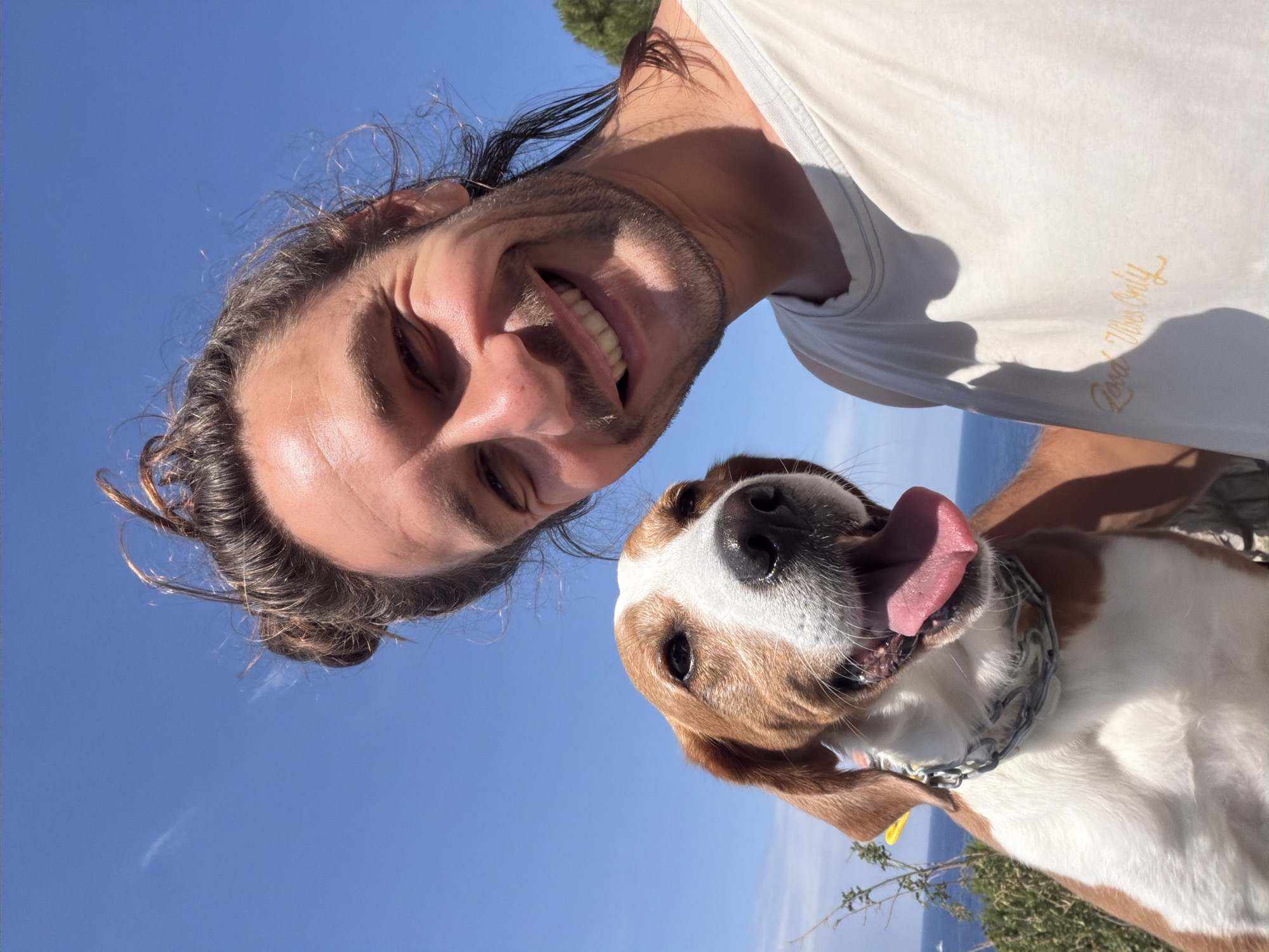This vibrant outdoor portrait features a man and a dog against a bright blue sky. The man, appearing to be in his 20s or 30s with long dark hair tied back and a beard, is positioned in the upper left, smiling broadly at the camera. He wears a white t-shirt with the golden cursive text "Good Vibes Only" visible on its right side. Below him and to the right, a medium-sized dog, with white and brown fur and resembling a Beagle mix, looks up at the camera with its tongue playfully hanging out. The dog is wearing a metal chain collar.
The image is captured from a low angle, highlighting the expansive, cloudless blue sky that fills much of the background. Strong, direct sunlight suggests it is daytime, likely morning or afternoon, with clear weather conditions. In the lower background, glimpses of a calm blue sea or ocean and some green foliage are visible, consistent with a coastal location like Mirište, Montenegro. Both the man and the dog appear joyful and content, sharing a happy moment outdoors.