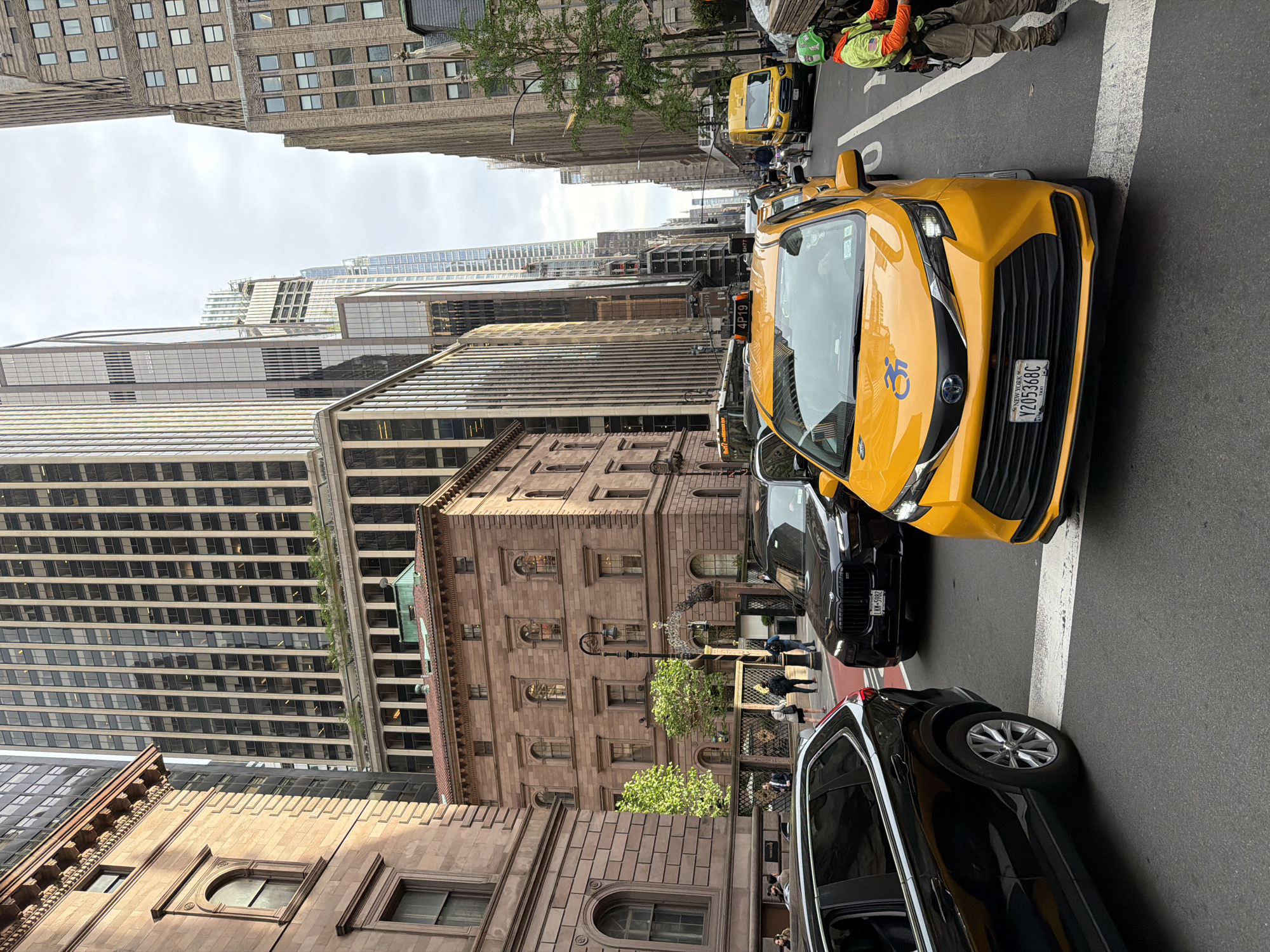 The image depicts a street scene in New York City. Several tall buildings are visible, constructed of stone and glass, indicating a dense urban environment. The sky is overcast. In the foreground, a yellow taxi is prominently positioned, with other black vehicles around it. A person wearing a high-visibility jacket and helmet is standing on the sidewalk to the upper right. There are other pedestrians on the sidewalk adjacent to a stone building with classical architectural details. The scene suggests a typical urban landscape with traffic and pedestrians in a business district.