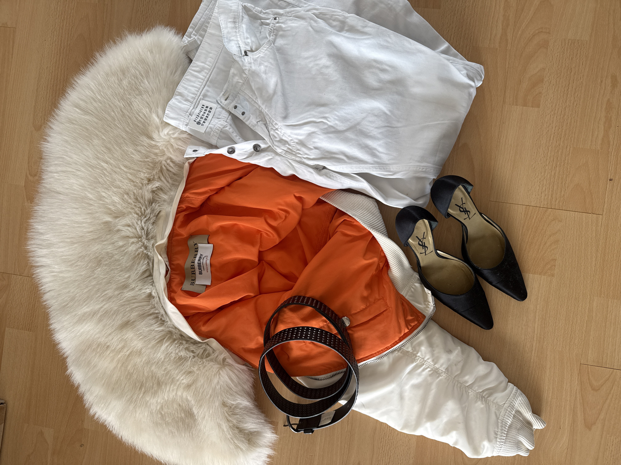 The image shows a still life arrangement of fashion items on a wooden floor.
Foreground items include a white bomber jacket with a faux fur hood and an orange lining, a black leather belt with studs, and a pair of black satin high-heeled shoes with a gold YSL logo. A pair of white trousers are folded on top of the jacket. The label on the jacket indicates that it is a Burberry item.
