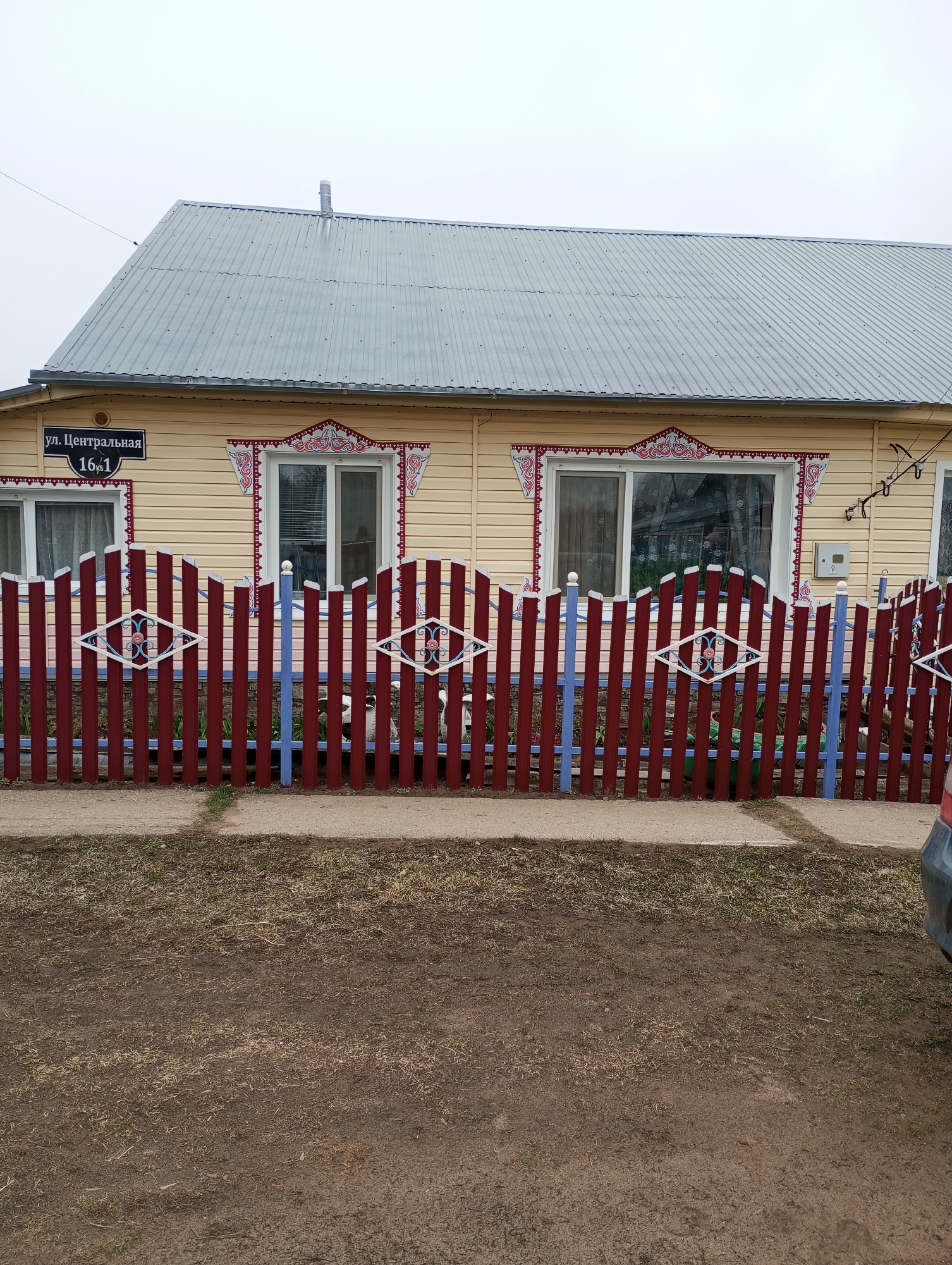 The image shows a house with a colorful fence. The house is one story with a metal roof and light yellow siding. Each window has detailed red and white trim. A sign reading "ул. Центральная 16к1" (Central Street 16/1) is visible on the corner of the house.
The fence in front of the house is made of alternating red and blue vertical boards, with white diamond-shaped decorations. A strip of concrete sidewalk runs along the base of the fence. The foreground is dirt and sparse grass. A portion of a car is visible on the right side of the image.
The location is Буренка, Russia, as inferred from the street address.