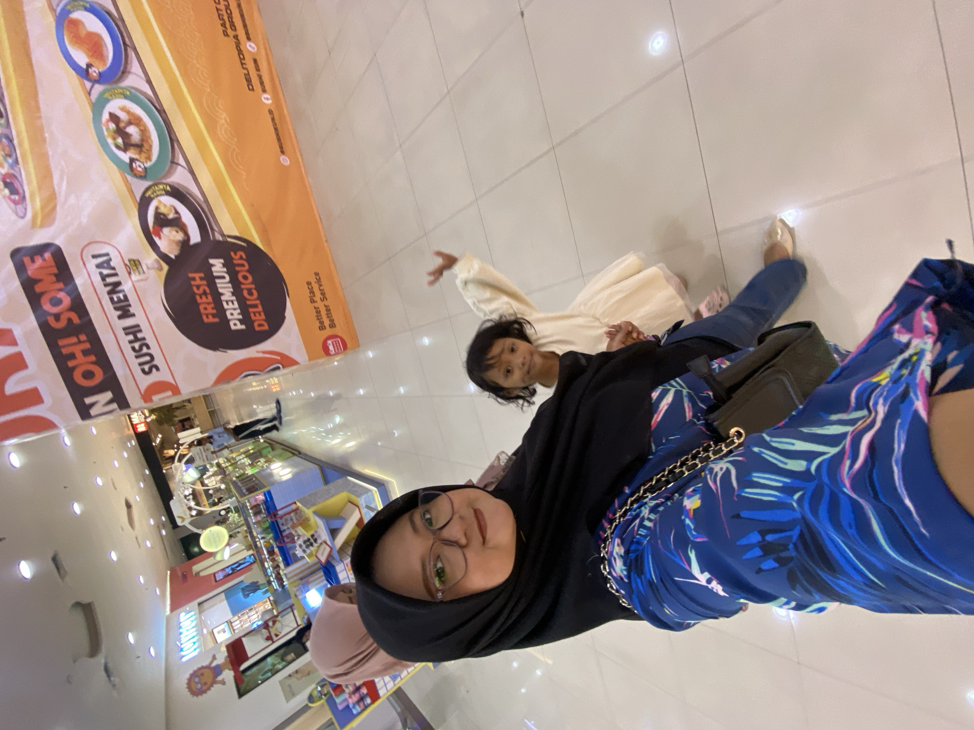 This image captures a selfie taken in a brightly lit indoor shopping mall, likely within the city of Sidoarjo, Indonesia. In the foreground, a woman wearing a black hijab and glasses is taking the photo, with a portion of her blue, tropical-patterned dress visible. To her right, a young girl in a cream-colored dress is looking towards the camera, her right arm raised. Partially visible behind the woman is another person wearing a pink hijab.
The background reveals a commercial environment with several shops. To the left, a store with bright signage and displays of merchandise is visible, featuring cartoon-like characters. Dominating the right side of the background is a large, orange banner advertising "OH! SOME SUSHI MENTAI," featuring images of sushi dishes. The floor of the mall is tiled with a light-colored material, reflecting the overhead lighting. The overall atmosphere suggests a casual outing in a retail setting.