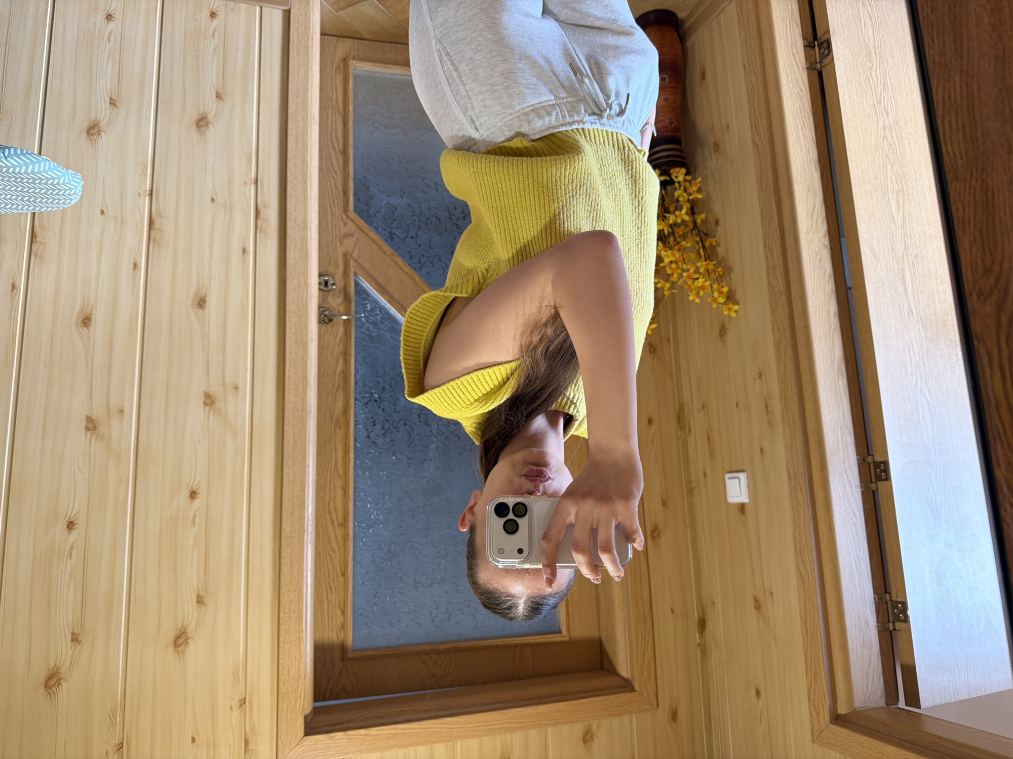 A woman takes a selfie indoors. She is wearing a yellow sweater and gray sweatpants. She is holding a white smartphone. The background shows wood-paneled walls, a door with a glass panel, and a vase with yellow flowers. There is a light switch on the wall. The location may be Buhovo, Bulgaria.