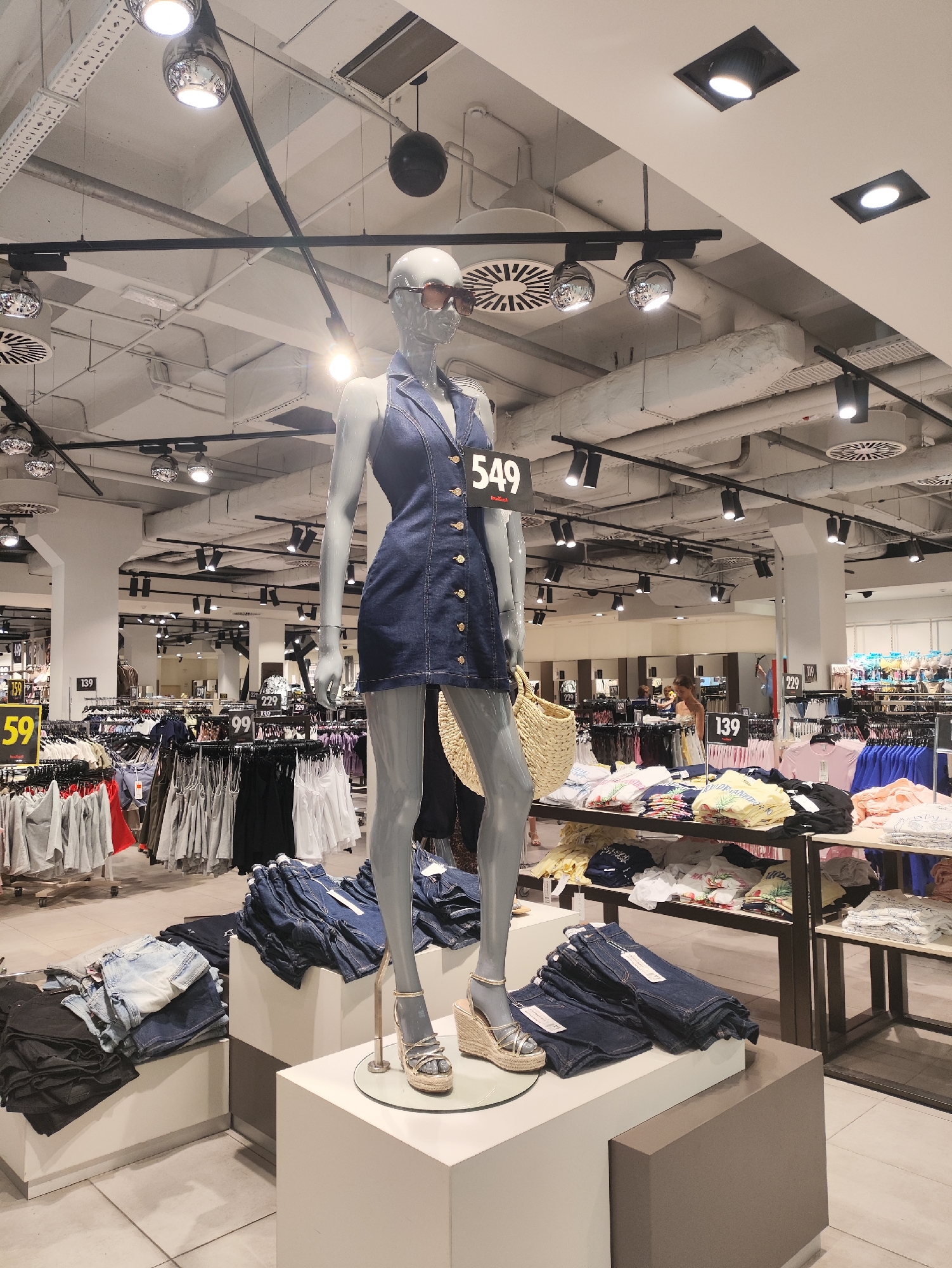 The image shows a retail store interior, likely a clothing store, in Chișinău, Moldova. In the foreground, a female mannequin is displayed wearing a dark blue denim dress and gold wedge sandals. The mannequin is positioned on a white pedestal. To the left of the mannequin, stacks of folded denim jeans are visible on a display unit. In the background, racks of clothing are organized on shelves and hanging displays. Several price tags are visible throughout the store, including "549", "159", "59", and "139". The store features industrial-style lighting with exposed pipes and ductwork on the ceiling. A few shoppers are visible in the background, browsing the merchandise.
