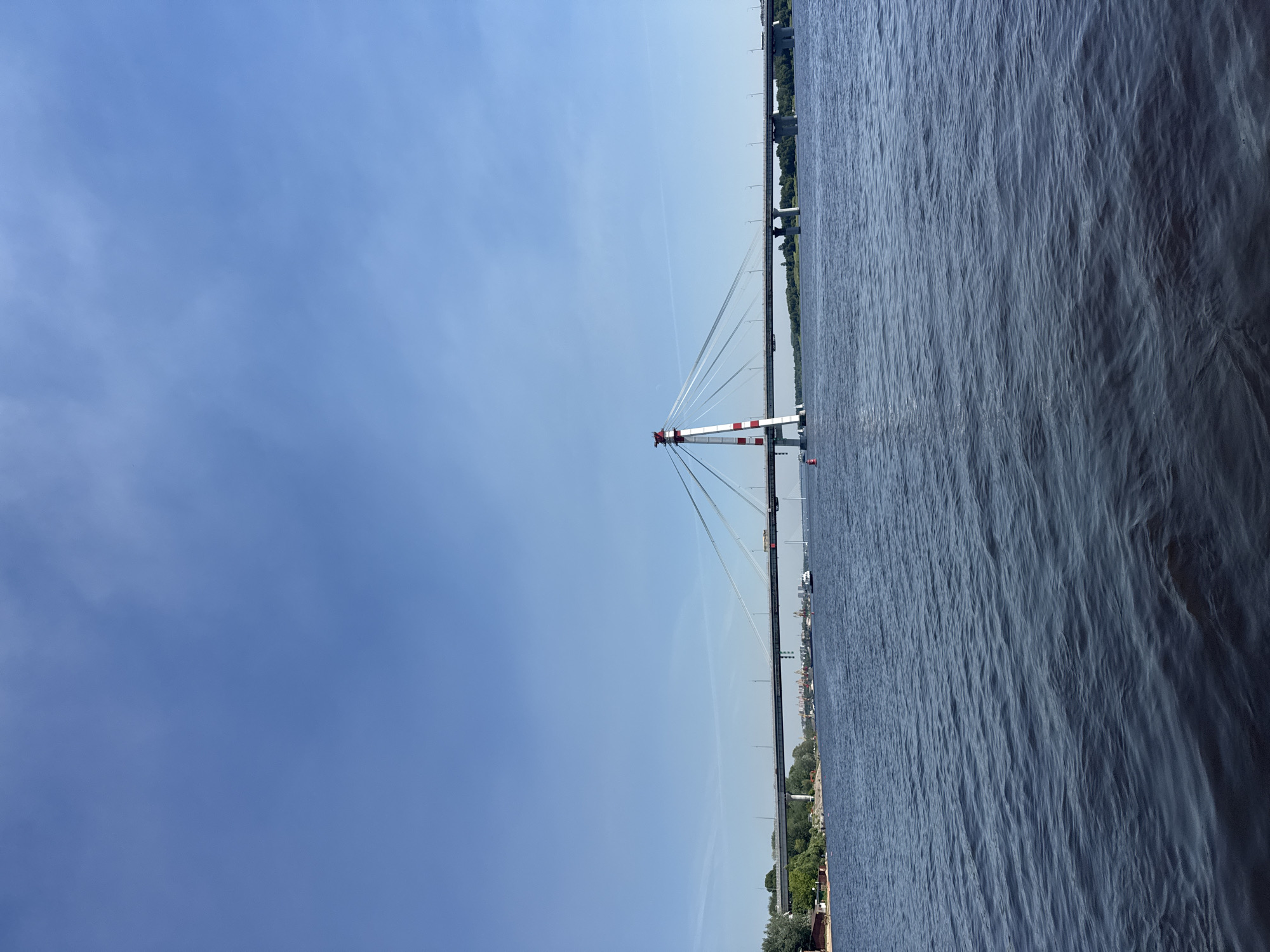 The image shows a cable-stayed bridge spanning a wide river under a clear blue sky. The bridge features a distinctive red and white striped tower supporting the cables. In the foreground, the dark blue water of the river is visible with moderate ripples. In the background, the far bank of the river is lined with trees and buildings, suggesting an urban or semi-urban environment. A few boats are present on the water, and vehicles can be seen on the bridge itself. A red buoy floats near the bridge tower. The overall scene depicts a sunny day with a focus on the bridge and its relationship with the waterway. This scene is likely the Severny Bridge over the Yagorba River in Cherepovets, Russia, given the distinctive bridge design and the geographical context of the Yagorba River flowing through the city. There are no people directly visible in the frame.