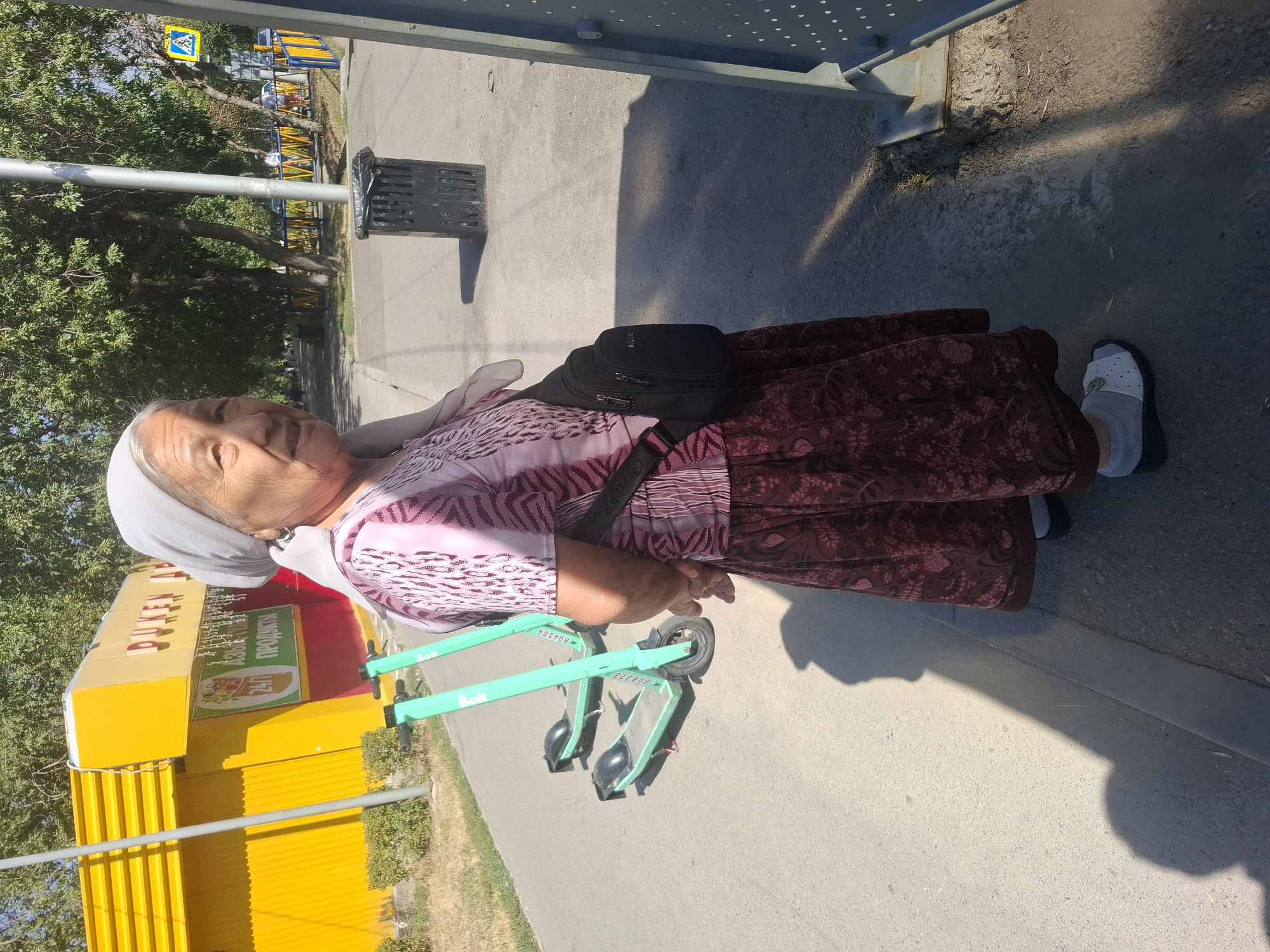 An elderly woman wearing a light gray headscarf, a patterned pink top, and a long dark red skirt with a floral print stands on a paved sidewalk. She is also wearing light gray socks and white flat shoes with a decorative element on the front. A black shoulder bag is slung across her body. Behind her, two green electric scooters are parked on the sidewalk. Further back, a bright yellow building with red trim has signage that includes "24/7 ПРОДУКТЫ" (24/7 PRODUCTS). The environment appears to be an urban setting with trees and a metal fence in the background. A black trash receptacle is visible to the right of the woman, and a pedestrian crossing sign is situated in the distance. The scene is illuminated by sunlight, casting distinct shadows. Based on the presence of "BOLT" scooters, the building signage in Russian, and the common urban landscape, this scene is likely located in Almaty, Kazakhstan.