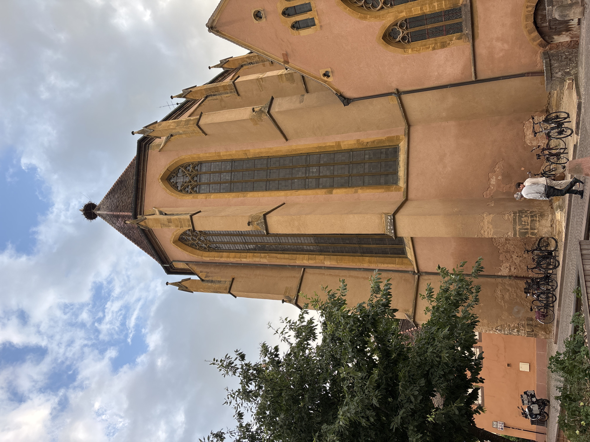 The image shows the Saint-Martin's Church, also known as the Collegiate Church of Saint-Martin, in Colmar, France. The church, with its characteristic salmon-pink facade, features tall, arched windows with intricate tracery. A prominent detail is a stork's nest atop the tiled roof. In the foreground, a tree with lush green foliage partially obscures the church. To the left, a motorcycle is parked on the cobblestone street. Several bicycles are leaned against the church wall. Two individuals, a man and a woman, are walking away from the camera on the paved pathway, with the woman appearing to be wearing a light-colored top and dark trousers, and the man in a light-colored top and dark trousers. The sky above is partly cloudy, with a mix of white and gray clouds against a blue background. The street is paved with cobblestones, and there is a low stone wall and some decorative planting beds. The overall scene depicts a quiet street in a historic European city.