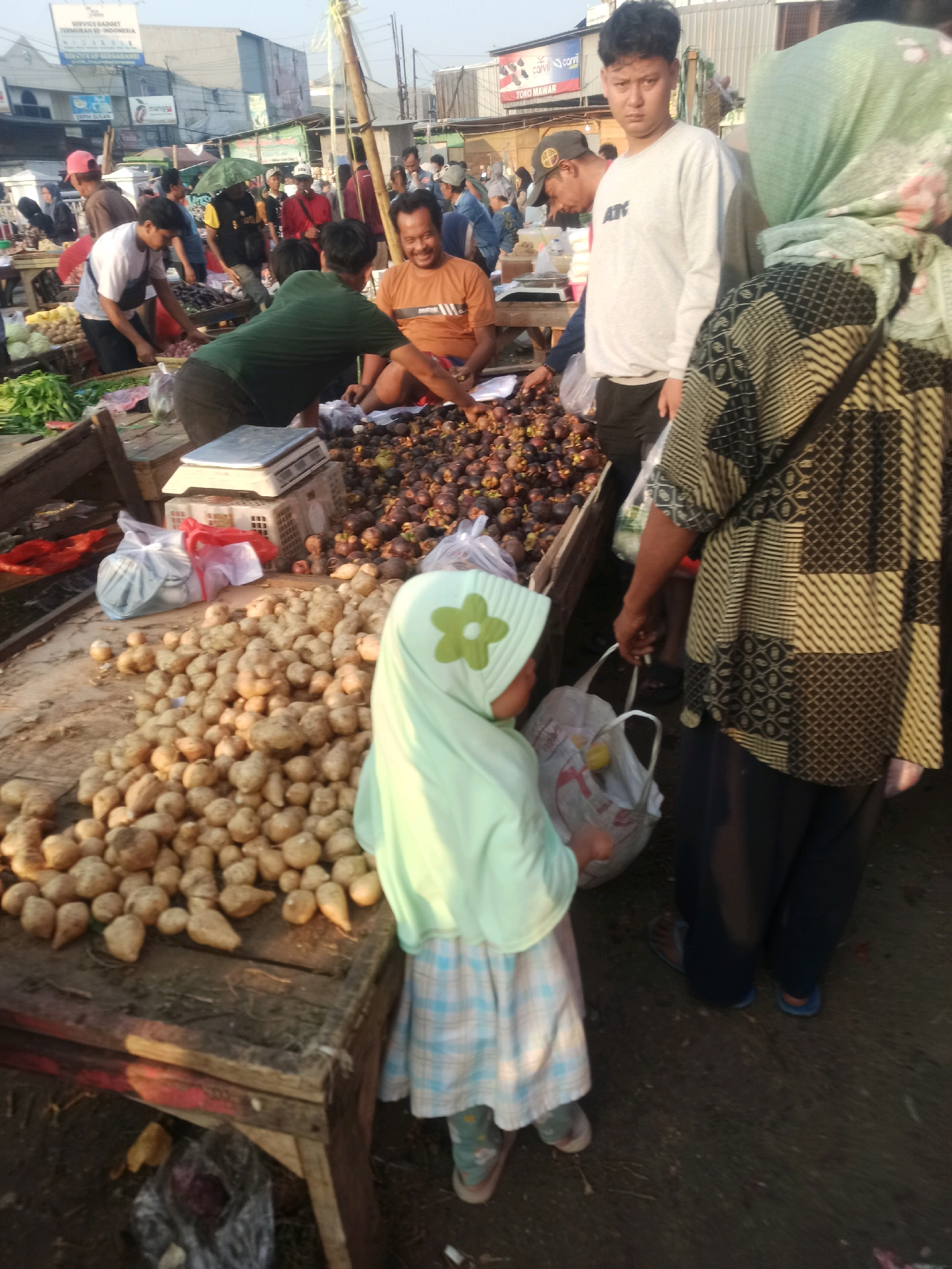 A street market scene in Cikarang, Indonesia. In the foreground, a young child wearing a light green hijab and a blue and white checkered dress stands facing away from the camera, looking at a display of root vegetables. To the right of the child, an adult wearing a patterned long-sleeved shirt and dark pants is holding a white shopping bag with a yellow object inside. Behind the child and adult, a stall is laden with mangosteens and other produce. Several other people are visible in the background, engaged in shopping or selling. The market appears to be outdoors, with stalls set up along a street or open area. The time of day is likely morning or late afternoon, as indicated by the warm lighting and long shadows.