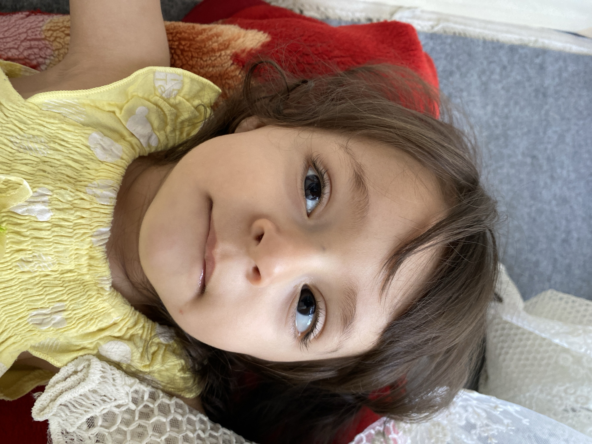 A close-up portrait shows a young child, likely female, lying down and looking directly at the camera. The child has fair skin, prominent dark brown eyes, and dark brown hair that falls around her face. Her expression is calm and gentle, with her lips slightly parted.
She is wearing a bright yellow top or dress featuring a textured, possibly smocked, design on the bodice, adorned with irregular white patterns that resemble polka dots. A portion of her left arm is visible in the upper part of the frame, and a section of white lace fabric, possibly from her clothing or a nearby item, is visible in the bottom right corner.
The child is resting on a soft surface. A warm-toned fabric, patterned in shades of red and orange, is visible beneath her head and shoulders, possibly a blanket or cushion. In the background, a gray carpet or rug can be partially seen, indicating an indoor setting. The lighting is soft and natural, emphasizing the child's features. No text is visible in the image. The context provided for the image is Kandahar, Afghanistan.