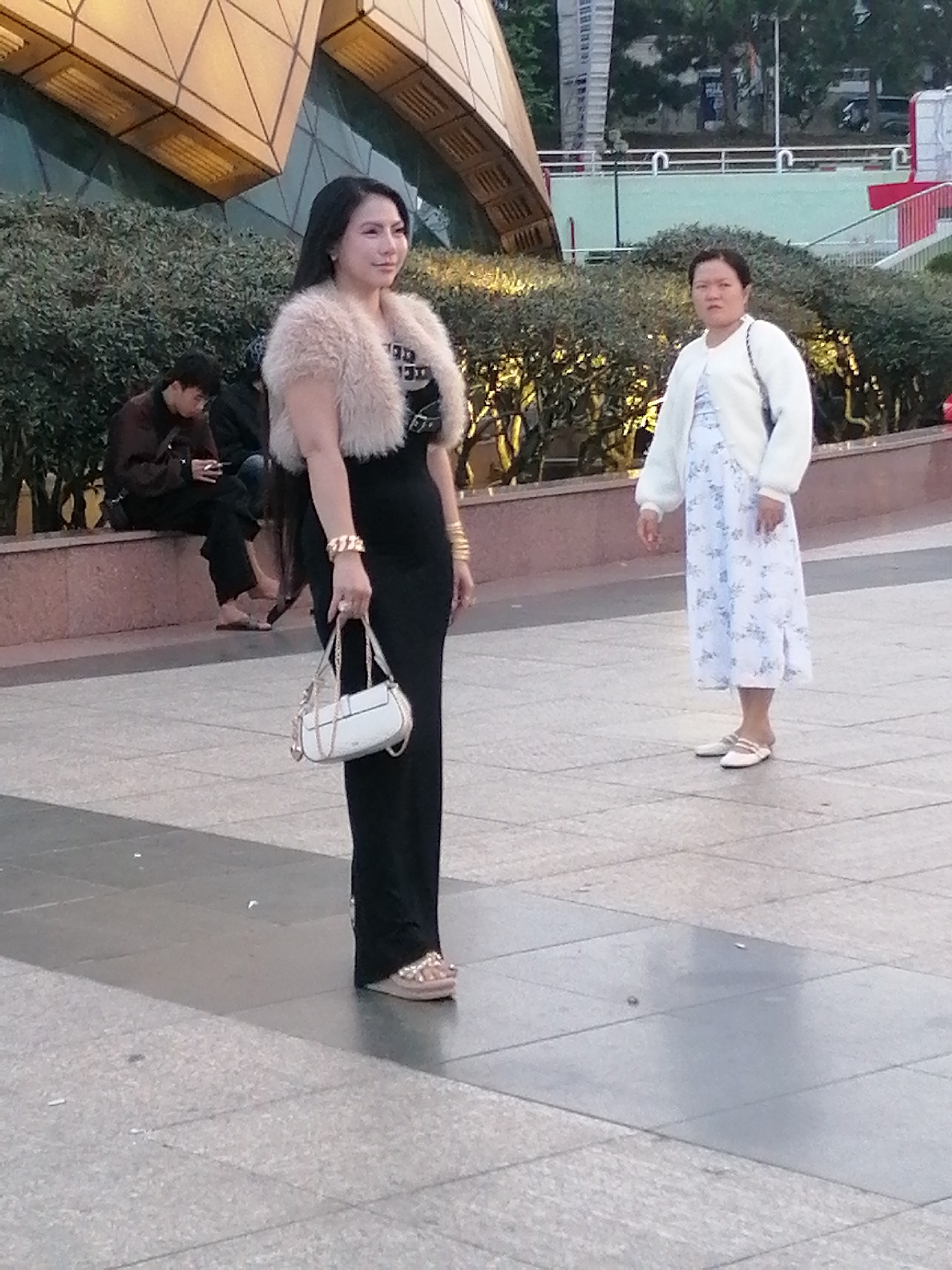 A woman stands in the foreground of the image, facing left, looking slightly away from the camera. She has long dark hair and is wearing a black dress, a fluffy beige vest, and gold jewelry on her wrist and hand. She is holding a white handbag with a chain strap. To the right of the woman, a second woman is standing and looking at the camera. She has short dark hair and is wearing a white cardigan over a light blue floral dress. In the background, a man sits on a ledge, looking down at his phone. The scene takes place on a paved outdoor area, likely a public space, with hedges and a modern building visible in the background. This scene is consistent with an urban environment in Da Lat, Vietnam, characterized by its architectural styles and public spaces.