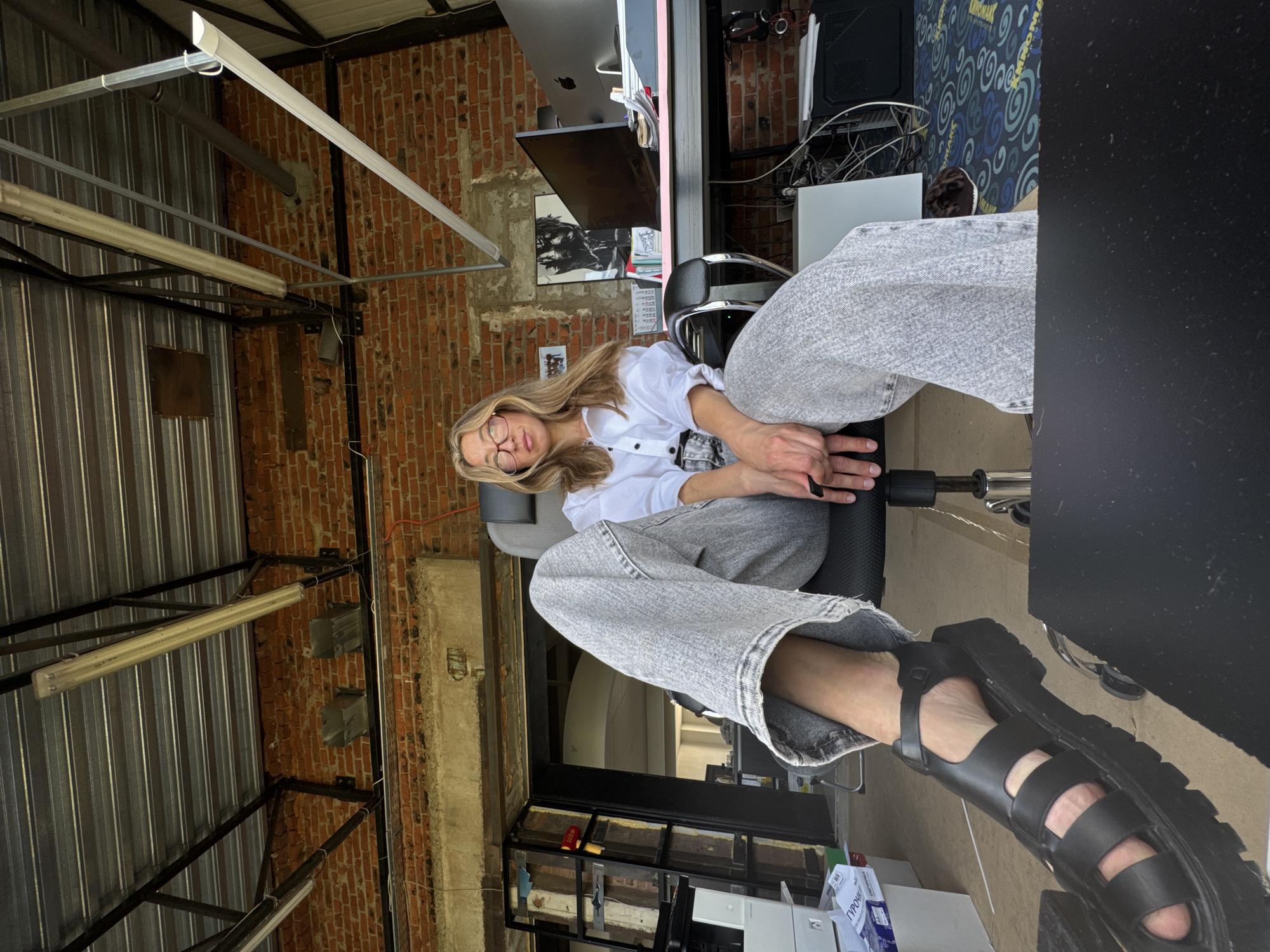 A woman with blonde hair and glasses is sitting in an office chair. She is wearing a white button-down shirt and wide-leg grey jeans. She has on black platform sandals. The office appears to be an industrial space with exposed brick walls and a metal ceiling. There is a desk with a computer monitor and other office supplies.