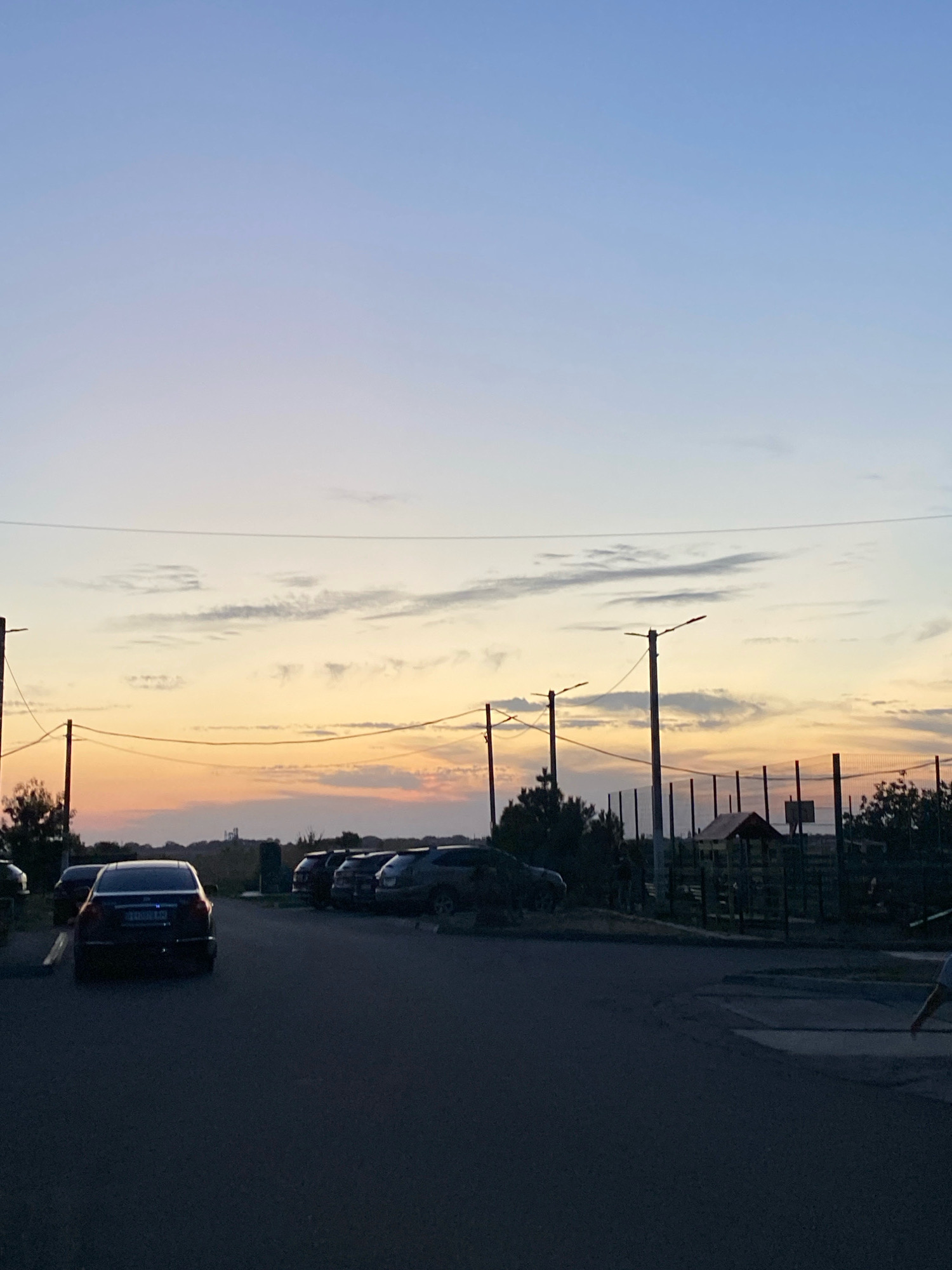 This image shows a parking lot at dusk with a clear sky transitioning from light blue to a pale orange horizon. In the foreground, a dark-colored sedan is parked, facing away from the camera. Several other vehicles are parked in a row behind it. A fence with a basketball hoop is visible in the background, along with some trees and a small structure. Power lines stretch across the sky. The scene appears to be in Chornomorsk, Ukraine.