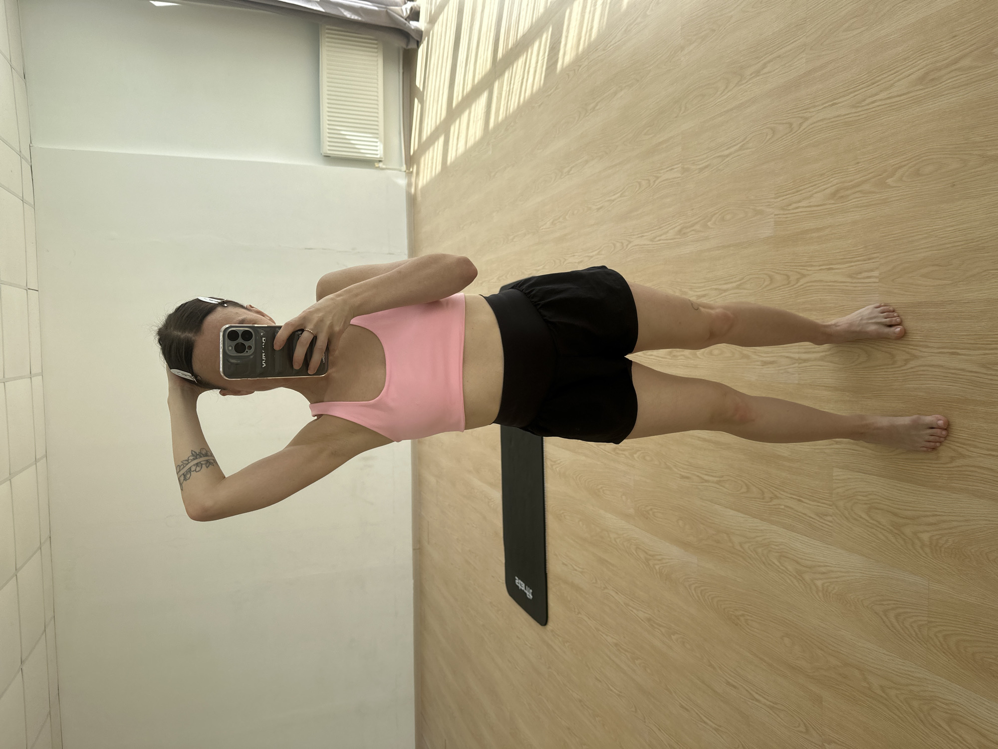 This mirror selfie, rotated 90 degrees clockwise, captures a person, likely female, in what appears to be an indoor exercise or changing room. The subject's face is obscured by a dark-colored smartphone, held to take the photo, which has a clear case with white letters "S.S" visible on the back. She is wearing a pink sports bra or crop top and black athletic shorts, and is barefoot. A band-style tattoo is visible on her left forearm, and a thin ring adorns her right hand.
The setting features a light-colored wooden floor with a distinct grain pattern, which occupies much of the lower frame. Prominent, parallel shadows, indicative of natural light streaming in from a window or opening, fall across the floor, suggesting it is daytime. The walls are light-colored, with a section of white tiles visible on the left and a white horizontal vent or grille in the upper left background. A black rectangular exercise mat, featuring mirrored white text (possibly a brand name), is laid on the wooden floor behind the person.
The attire and the presence of an exercise mat suggest the person might be in a gym, yoga studio, or a home workout space in Votkinsk, Russia.