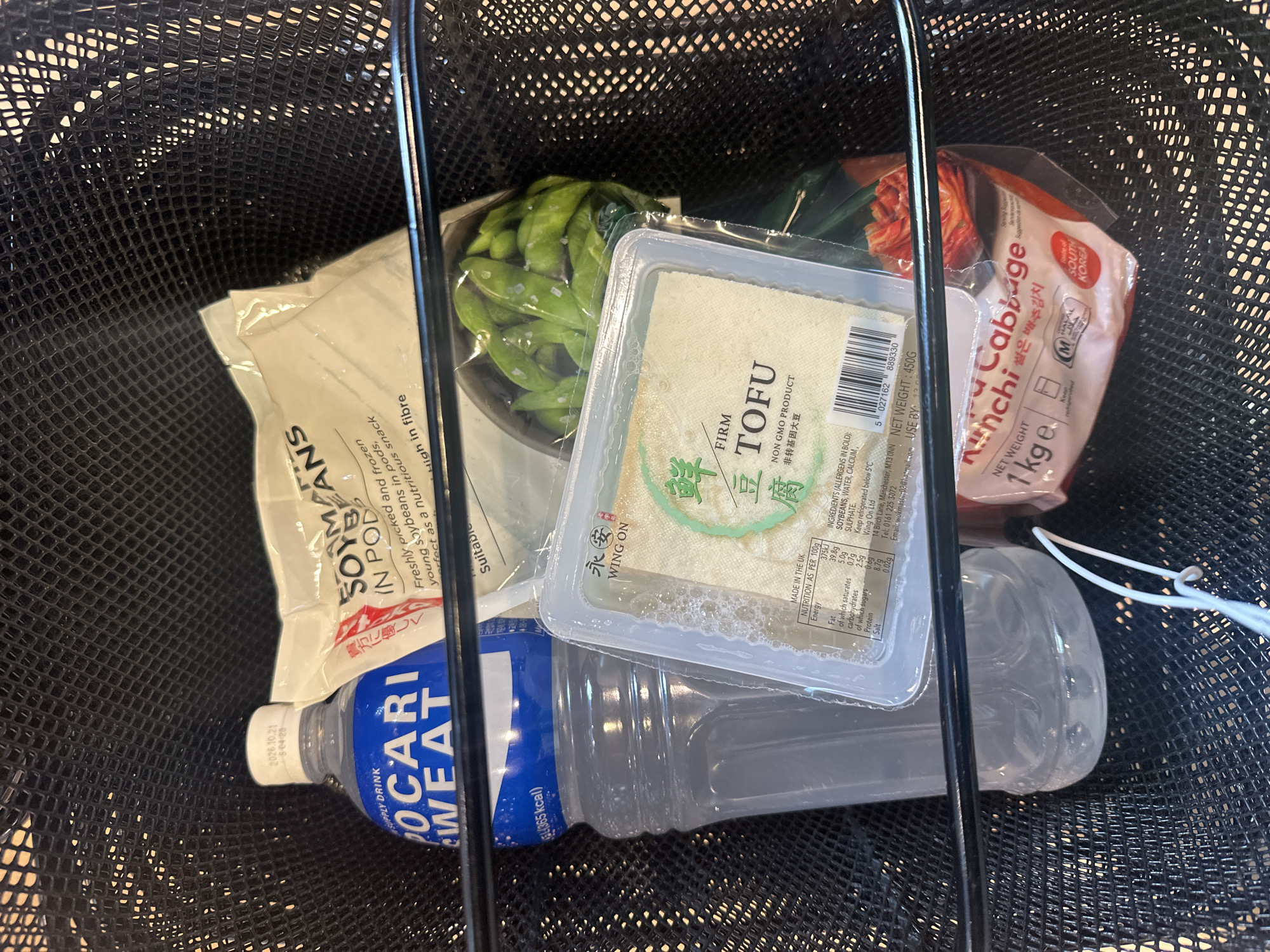 A black mesh shopping basket contains groceries. The items include a bag of frozen soybeans in pods, a container of fresh edamame, a package of firm tofu produced by Wing On, a bottle of Pocari Sweat sports drink, and a 1 kg package of kimchi made in South Korea. The packaging on the tofu indicates that the product was made in the UK and lists an address in Manchester. The location is not in London, United Kingdom.