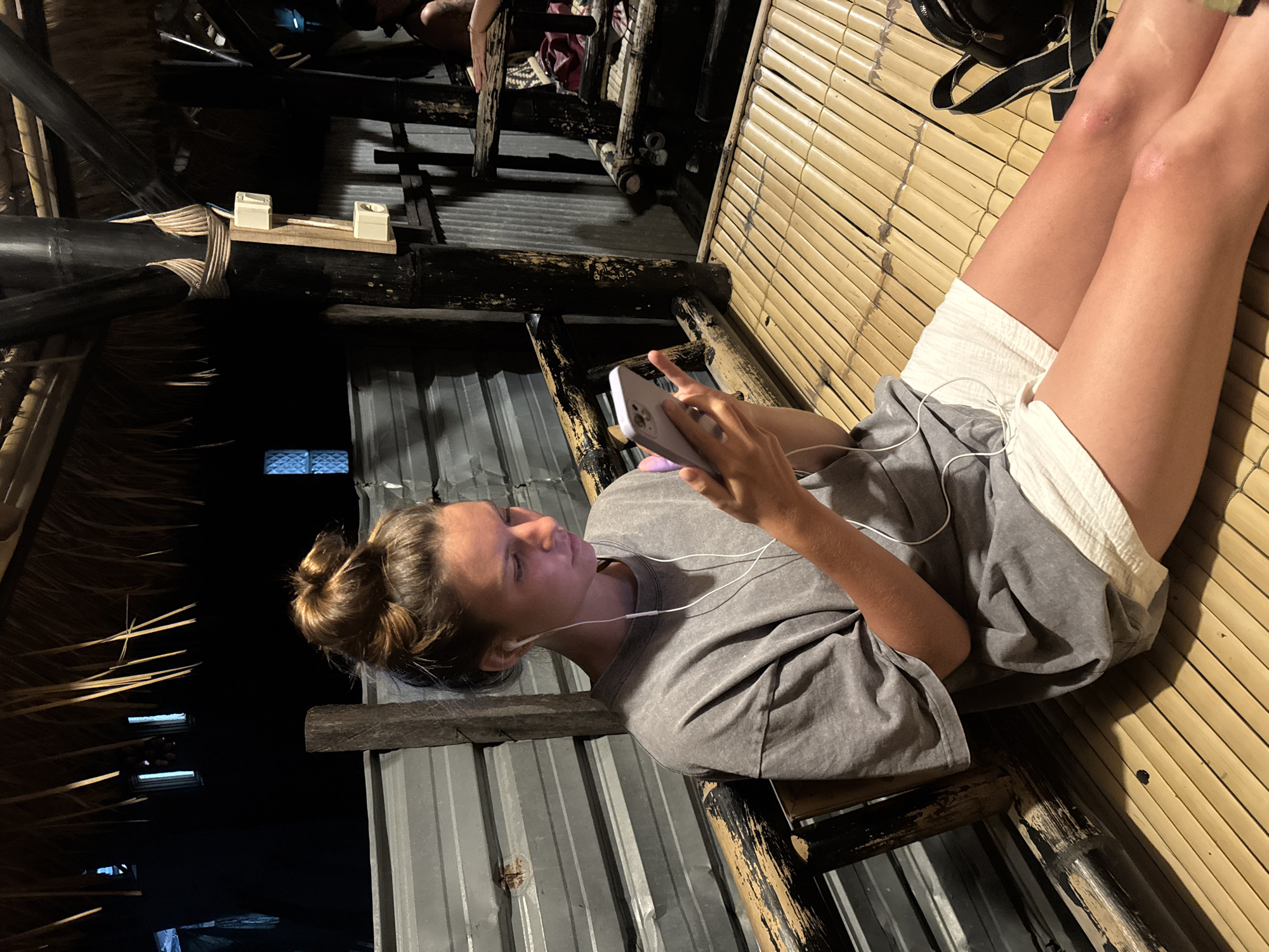 A young woman with her hair in a bun, wearing a grey t-shirt and white shorts, sits on a bamboo mat floor. She is holding a light purple smartphone and appears to be looking at it while wearing white earbuds. The background consists of a corrugated metal wall and a thatched roof supported by bamboo poles. The scene is dimly lit. The location is a communal outdoor seating area, possibly a restaurant or rest stop, in Kertasari, Indonesia.