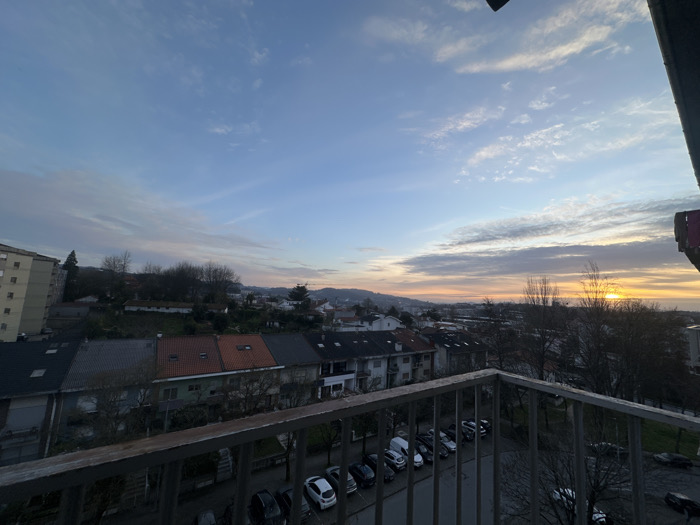The image presents an elevated, wide-angle view of a residential urban landscape in Guimarães, Portugal, observed during either sunrise or sunset. The immediate foreground is dominated by a dark, weathered metal balcony railing composed of a horizontal top bar, vertical balusters, and a lower horizontal support.
Beyond the railing, a street stretches horizontally across the midground, occupied by numerous parked vehicles including various sedans, hatchbacks, and a white commercial van. The street surface appears dark. To the left, a multi-story apartment building with a light-colored facade is partially visible. Further into the midground, a row of lower-rise residential buildings is visible, distinguished by roofs of varying colors, including dark grey, reddish-brown, and light green.
Scattered throughout the scene are numerous deciduous trees, mostly bare of leaves, indicating an autumn or winter season. In the background, distant hills or elevated terrain are visible, blending with the horizon. The sky transitions from a pale blue overhead to warm orange and yellow tones towards the right horizon, where a bright, circular solar disk is present, partially obscured by tree branches and distant structures. Wispy, elongated clouds are distributed across the sky. No individuals are visible within the depicted scene.