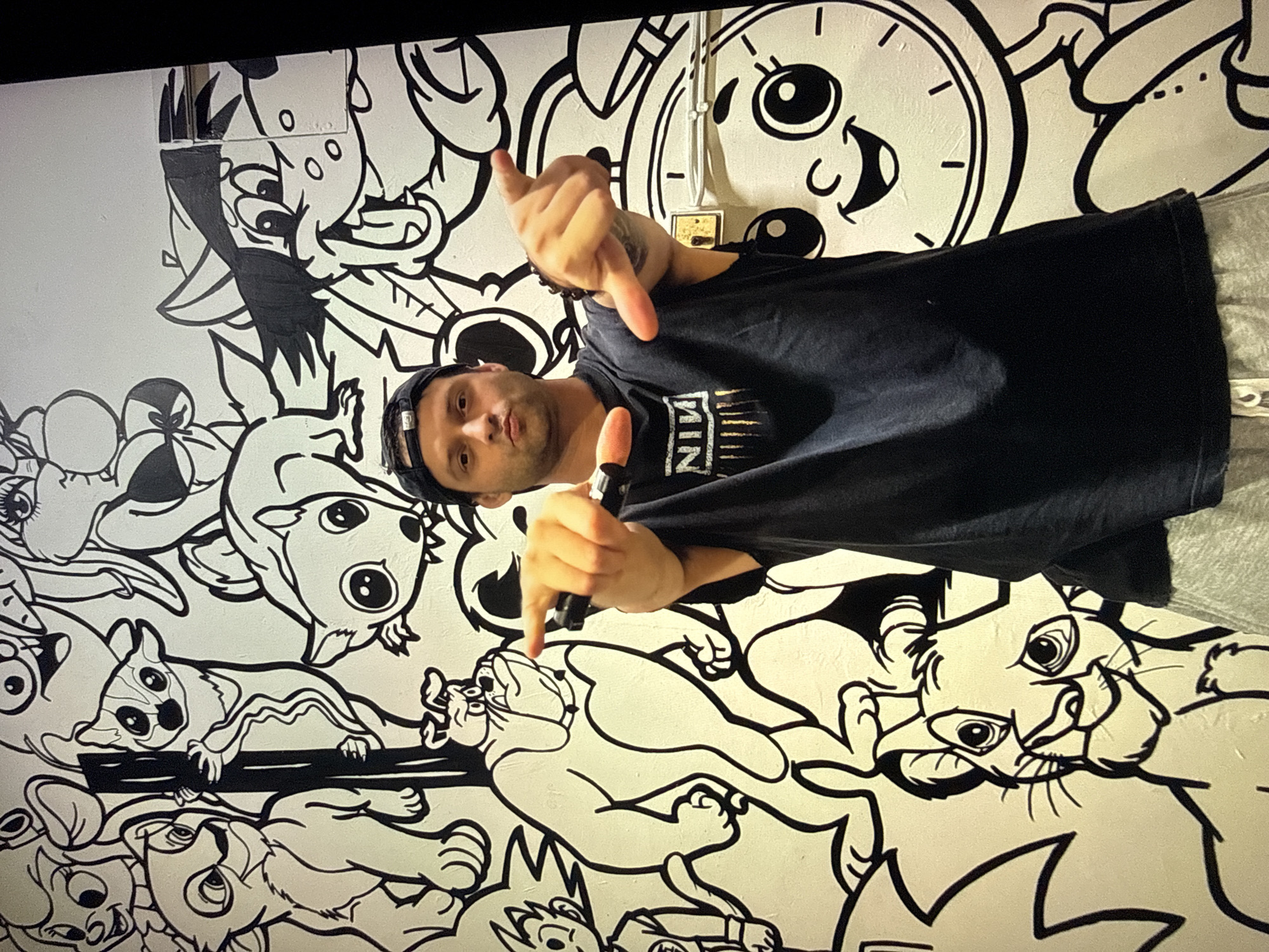 In a dimly lit room, a man wearing a black t-shirt with "NIN" emblazoned on it and a black baseball cap stands in front of a black and white mural. The mural features a variety of cartoon characters, including figures reminiscent of "The Lion King" and other animated animals. The man is holding two black objects, possibly markers or pens, in his hands. His right hand is extended forward, with his index finger pointing towards the camera, and his left hand is similarly extended. He is looking directly at the camera with a neutral expression. The environment appears to be an indoor space with a large mural covering the wall behind the man. The mural includes a stylized clock face with a smiling face. The location is identified as Taipei, Taiwan.