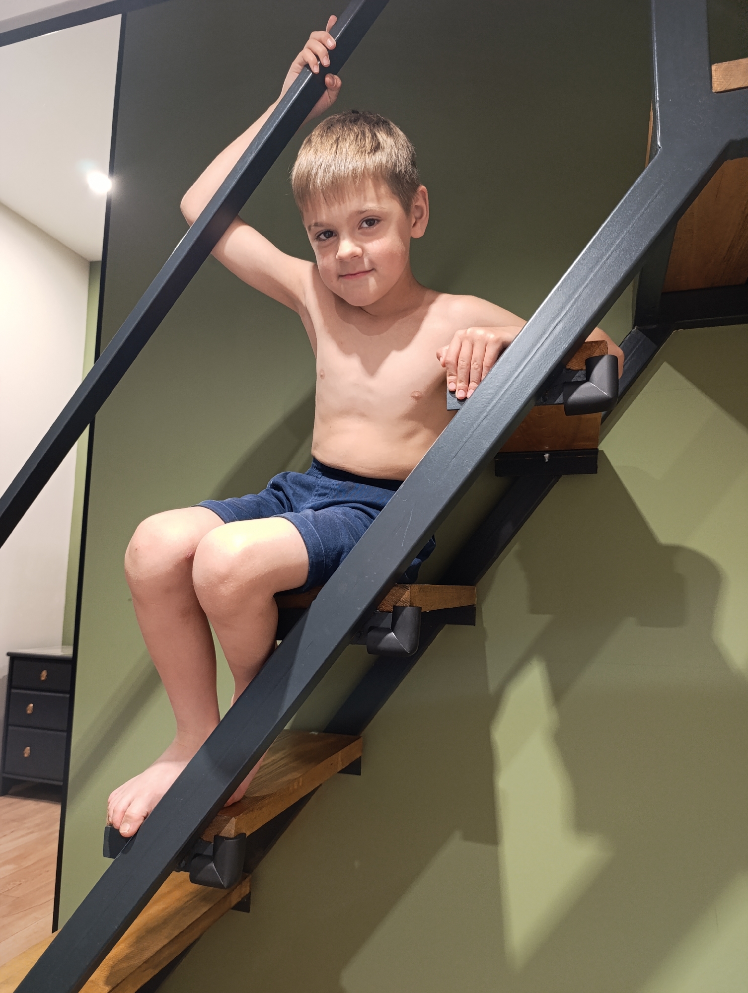 A young boy, appearing to be between 6 and 10 years old, is sitting on a modern, open-tread staircase indoors. He has short, light brown hair and fair skin, and is shirtless, wearing dark blue shorts. The boy looks directly at the camera with a gentle, friendly smile. His left hand grips the dark grey metal frame of the staircase, while his right arm rests casually on a lower section.
The staircase features a robust dark grey metal frame with light wooden treads. Black L-shaped corner protectors are visible on some of the treads. The wall behind the staircase is painted a muted olive green. Artificial lighting illuminates the scene, creating soft shadows on the wall, particularly to the right. In the upper left background, a light fixture can be seen, and a partial view of a black dresser with brass-colored handles is visible in what appears to be an adjacent space or reflection. The floor in this area is light-colored wood. The setting suggests a contemporary home or apartment in Moscow, Russia. The boy's expression conveys a sense of comfort and ease. No text is visible in the image.