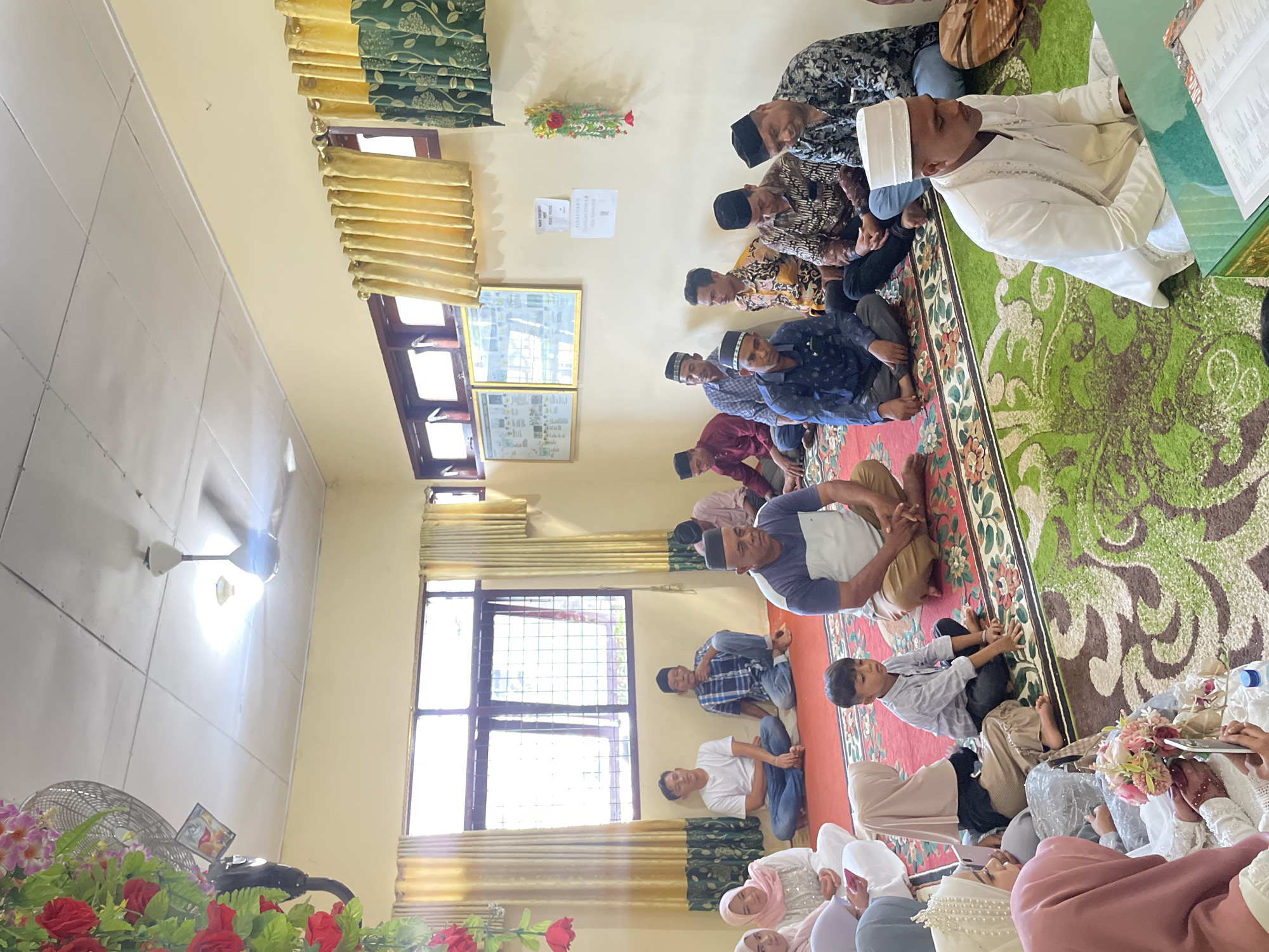 In an indoor room, likely in Pasar Paloh, Indonesia, a group of people are gathered, seated on the floor on a large, ornate rug. The room has light yellow walls and is illuminated by ceiling lights and natural light from windows covered by yellow and green patterned curtains. In the foreground, a man in a white traditional outfit with a white cap is seated on the floor, facing slightly to the right, looking down. To his left, a young boy in a light blue shirt is also seated, looking forward. To the left of the boy, several women are seated, some wearing hijabs in pink and beige. One woman in a white wedding dress with a beaded headpiece is holding a bouquet of flowers and looking at her phone. Further back, a group of men are seated, all wearing traditional attire and many with caps. One man in a white t-shirt and blue jeans is seated cross-legged. A ceiling fan is visible in the upper left corner, along with decorative red flowers. Several framed posters or pictures are on the wall in the background. The overall atmosphere appears to be a formal gathering or ceremony.