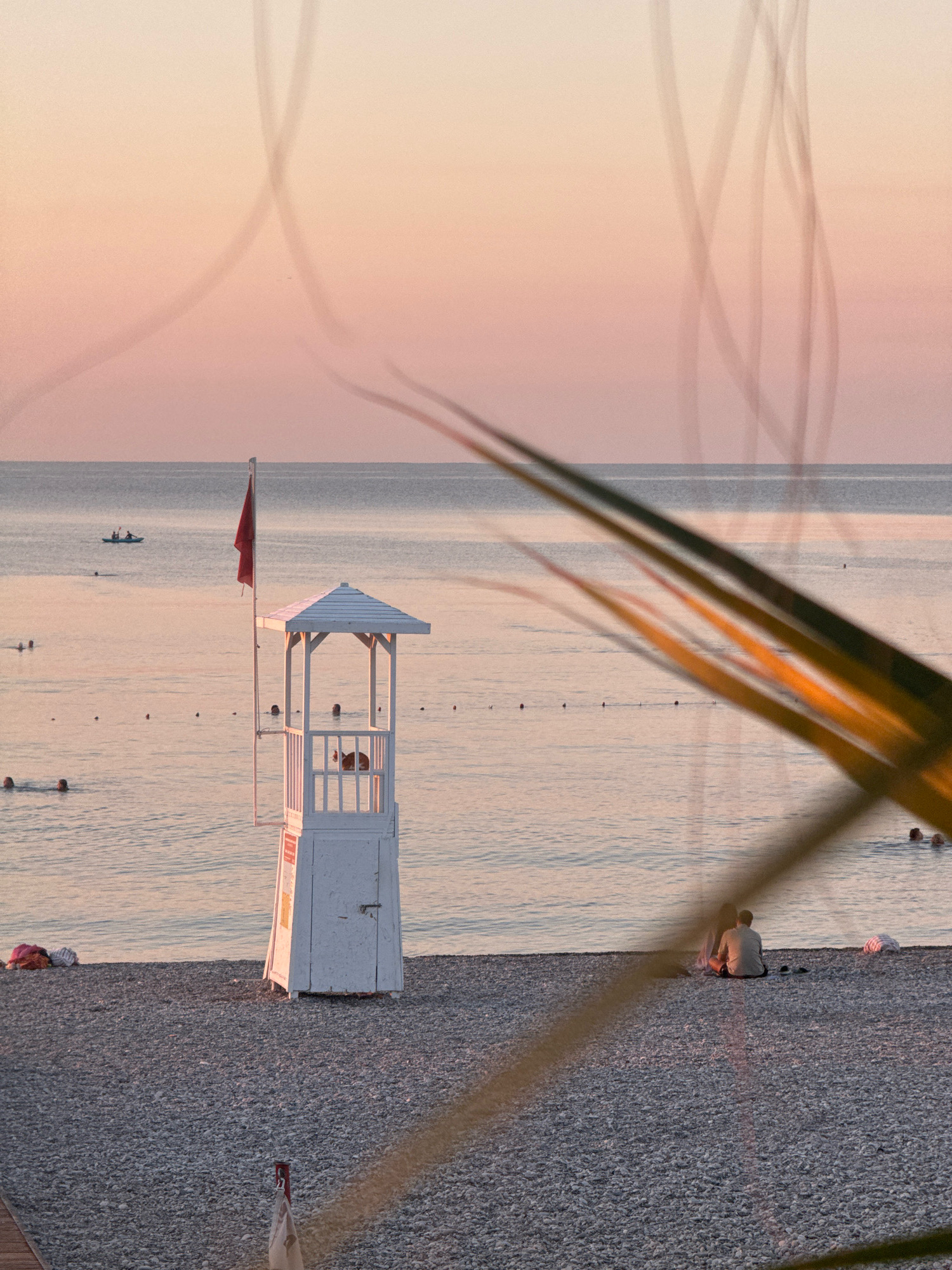 The image depicts a serene beach scene in Antalya, Turkey, during sunset. The sky transitions from a soft pink to a pale orange, with faint contrails from an aircraft visible. In the foreground, a white lifeguard tower stands prominently on a pebbled beach. A red flag flies from the tower. Several people are visible in the water, some swimming and others in a tandem kayak in the distance. On the beach, a couple sits together, silhouetted against the calm sea. The image is partially obscured by out-of-focus palm fronds in the foreground, adding depth and a tropical feel to the composition. The lighting suggests the golden hour, casting a warm glow over the entire scene.