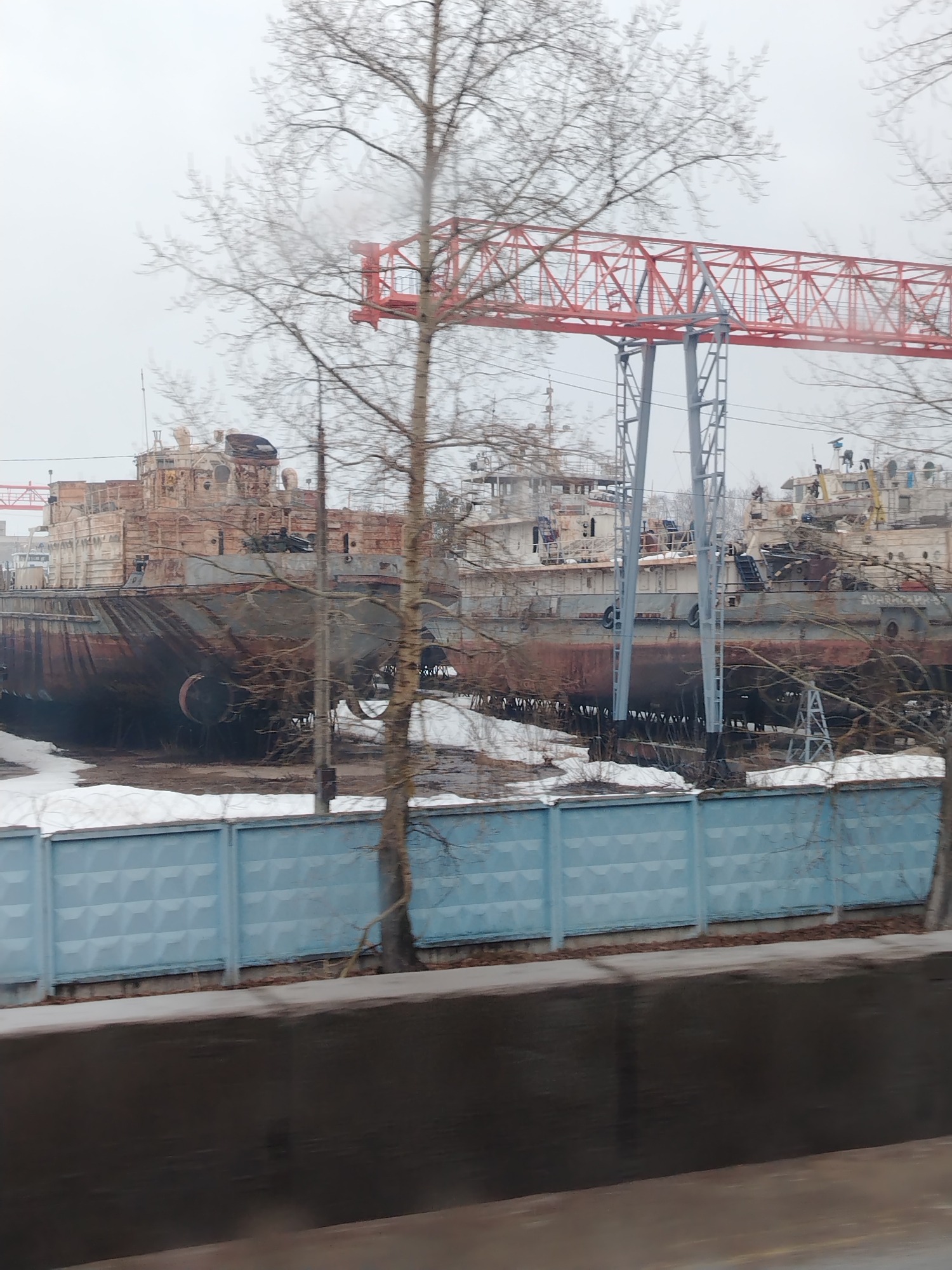The image depicts a shipyard in what appears to be the city of Chaikovsky, Russia. Several large, aging vessels are visible, seemingly in a state of disrepair. A prominent red gantry crane looms over the ships. In the foreground, a light blue concrete fence partially obscures the view of the shipyard. Patches of melting snow are present near the fence and around the ships, indicating a cold, possibly early spring, environment. Bare trees are scattered throughout the scene, typical of the season. The sky is overcast, contributing to a muted color palette. The image is taken from a lower angle, possibly from a moving vehicle, and focused on the ships and industrial infrastructure.