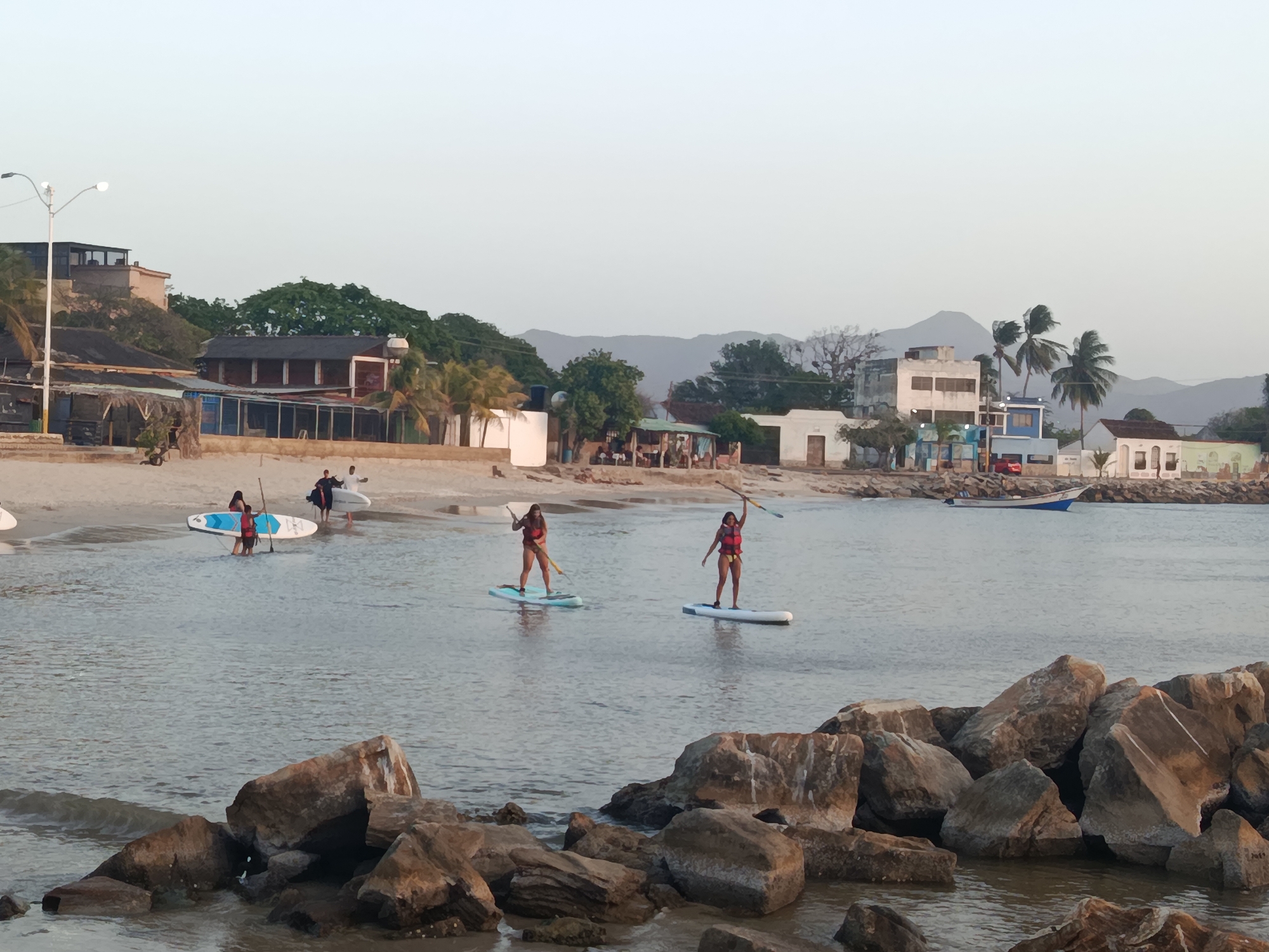 The image depicts a coastal scene in Juan Griego, Venezuela, featuring people engaged in paddleboarding. In the foreground, a rocky outcrop extends into the water. Two women, wearing life vests, are actively paddleboarding on the calm water. Further back, on the beach, several individuals are carrying surfboards. The background includes buildings typical of the town, blending with the natural landscape of trees and distant hills under a clear, pale sky.