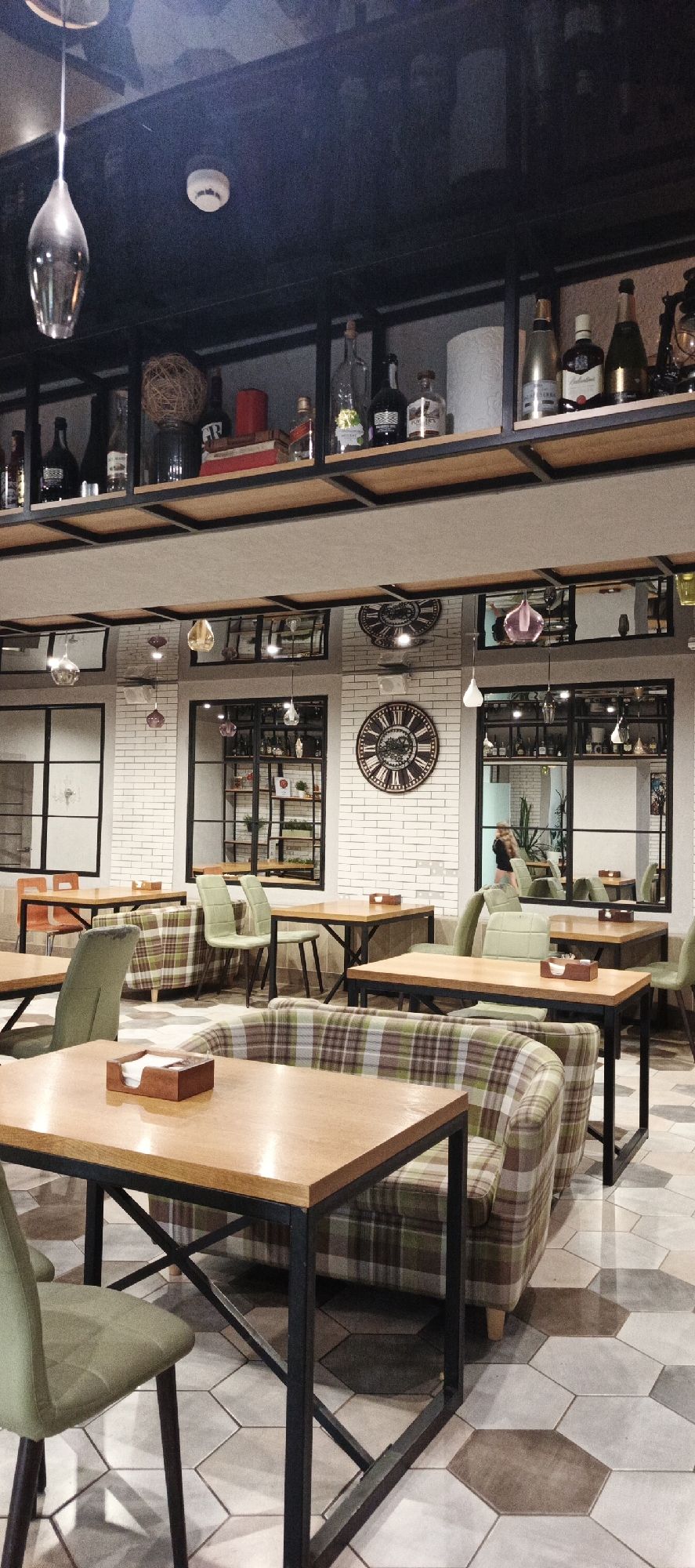 This is a medium shot, eye-level, interior photograph of a restaurant dining area. The scene is set during what appears to be nighttime, as evidenced by the artificial lighting. The restaurant has a modern industrial aesthetic, with exposed shelving units filled with bottles and decorative items, metal-framed tables, and chairs upholstered in a green and brown plaid fabric. The walls are white brick, and the floor is tiled with large, hexagonal tiles in varying shades of gray and brown.
Prominently featured are several tables set for dining, some with chairs pulled up, suggesting a quiet evening service. The overall atmosphere appears calm and inviting. Notable details include a large, ornate clock on the back wall, reflecting in a mirror above, and several pendant light fixtures hanging from the ceiling. The shelves above the seating area display a collection of various bottles, some appearing to be alcohol, as well as decorative items like a woven ball and books. In the distance, through a mirrored panel, a person can be seen standing. There is no visible text in the image. The setting suggests a restaurant or bar in Votkinsk, Russia.