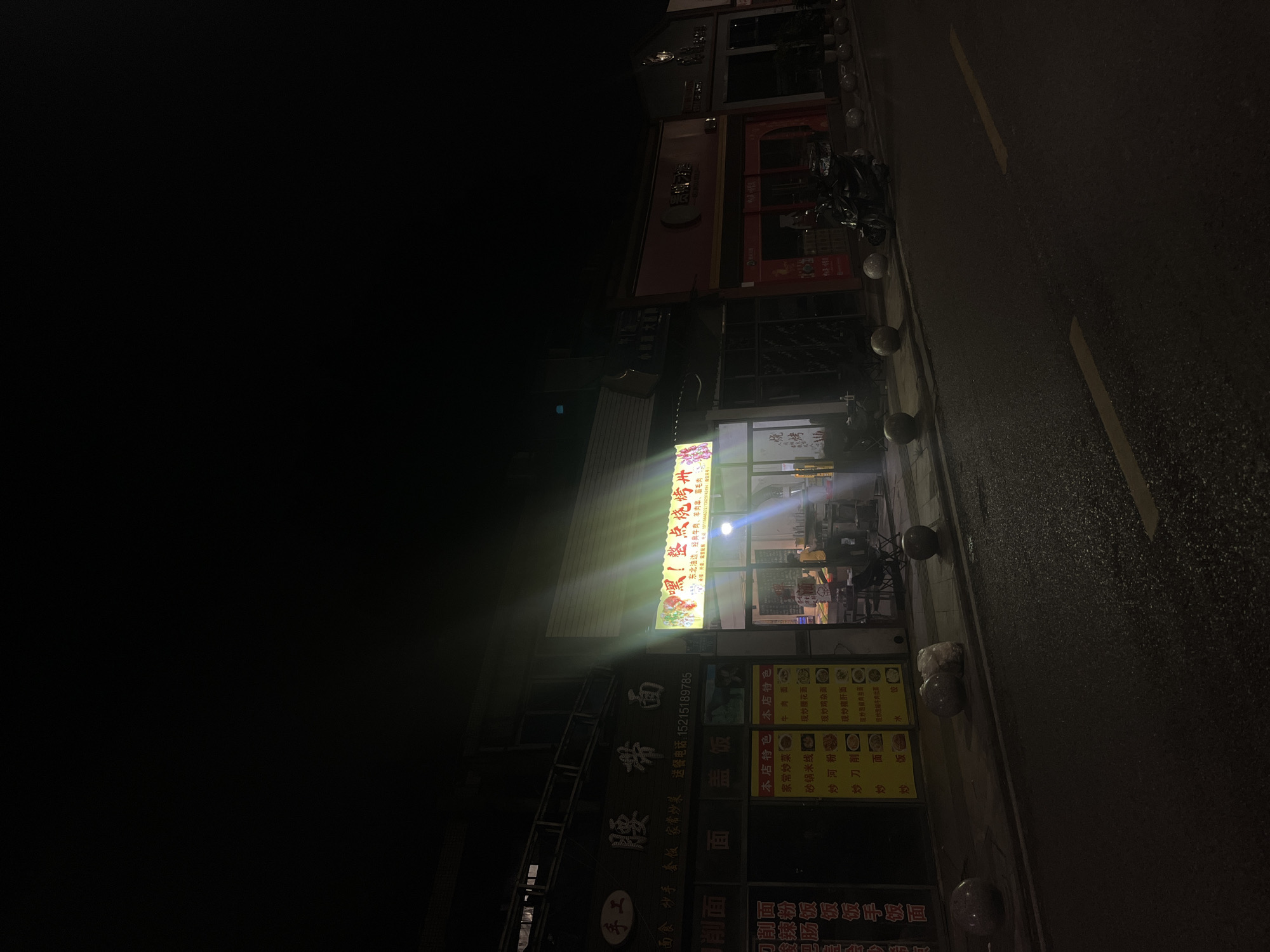 This is a nighttime street scene in Jiulongpo District, China, featuring a row of commercial buildings illuminated by storefront lights. On the left, a "手工 腰带面" (Handmade Belt Noodles) shop is visible, displaying menu items with images and a phone number (15215189785). To its right, a brightly lit storefront features a large sign reading "嘿! 整点烧烤 烤串" (Hey! Let's get some BBQ Skewers), with additional text specifying "东北油边, 经典牛皮, 牛肉串, 羊毛串" (Northeastern oil-side pork, classic beef skin, beef skewers, lamb skewers) and two phone numbers (15156640570, 13355721624). Inside this shop, two indistinct figures are visible; one appears to be standing behind a counter, and another is seated at a table. Further to the right are darker storefronts, one with a red facade and another with a dark facade, where a black scooter is parked on the sidewalk. The asphalt road in the foreground shows a yellow dashed line on its right side and appears somewhat reflective. Multiple spherical stone bollards of varying colors are spaced along the sidewalk, with a white bag of litter situated next to one bollard. The sky above is dark.