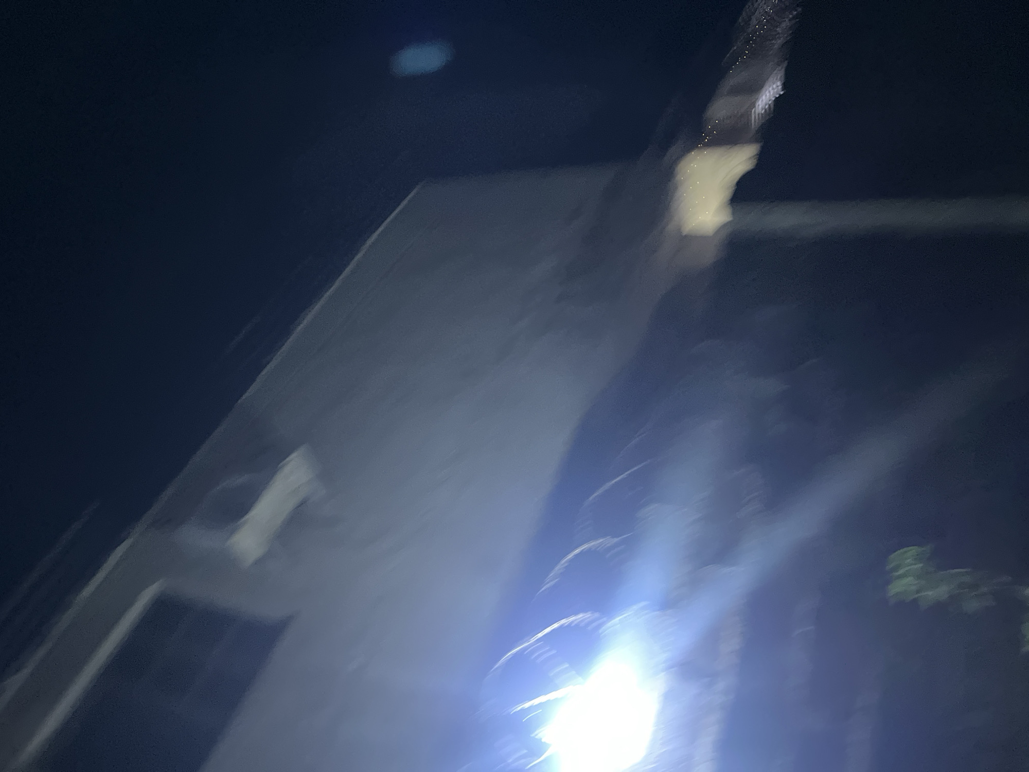 The image shows a blurred view of a building at night. The building appears to be made of concrete, with a few visible windows. A bright light source is present near the bottom of the frame, causing significant glare and obscuring details. Some trees are also visible on the right side. The exact location in Abuja, Nigeria is indeterminable from the image.