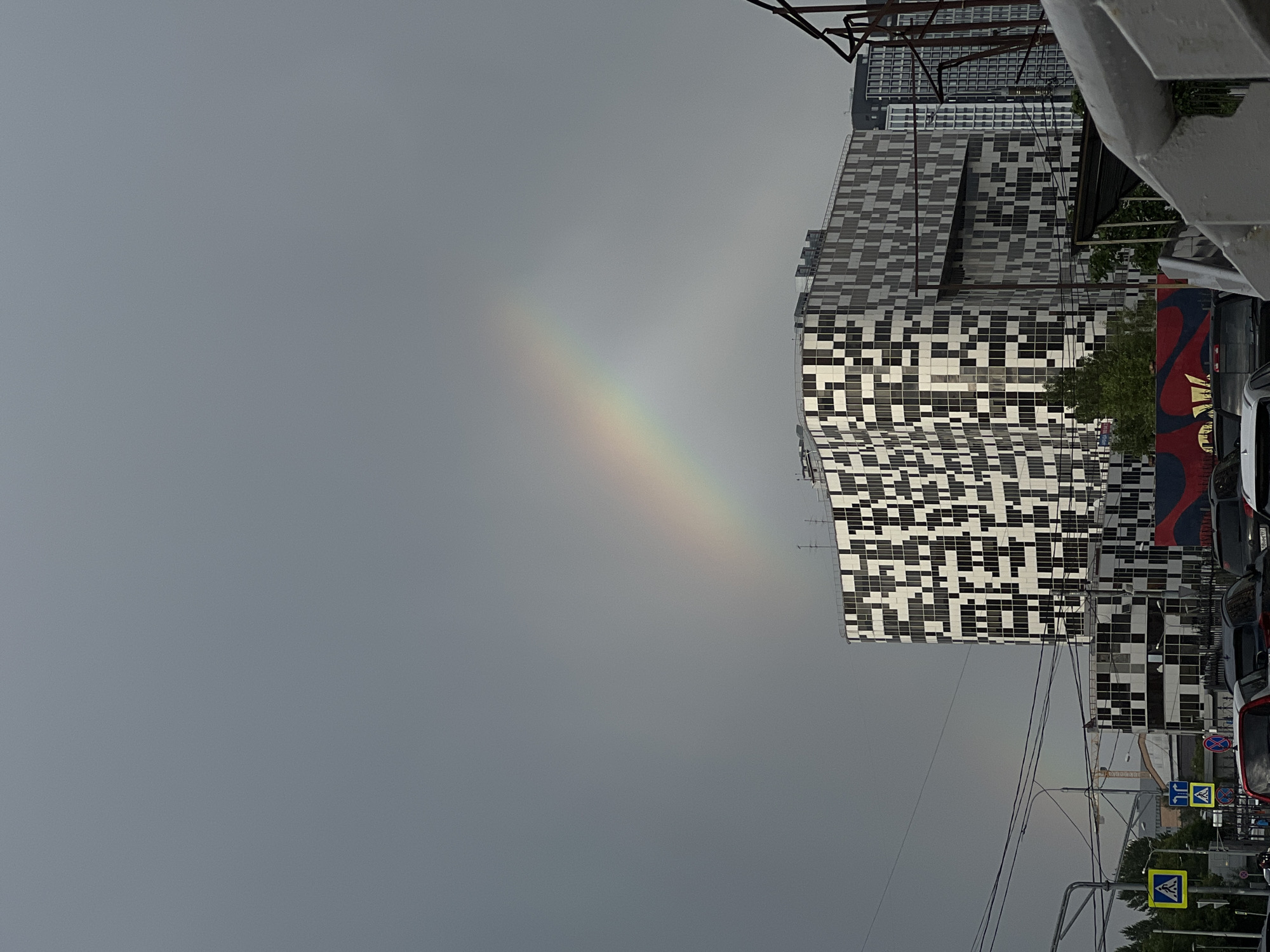 The image depicts a cloudy sky with a faint rainbow visible. In the foreground, a modern building with a striking black and white checkered facade stands tall. To the right, a portion of a white structure, possibly a wall or building, is visible. Electrical wires crisscross the scene, and street signs are present, including a pedestrian crossing sign with a yellow background and a silhouette of a person walking. Cars are parked on the right side of the image. In the background, more buildings can be seen, including a taller structure with a grid-like pattern on its facade. The overall impression is an urban setting on a day with unsettled weather.